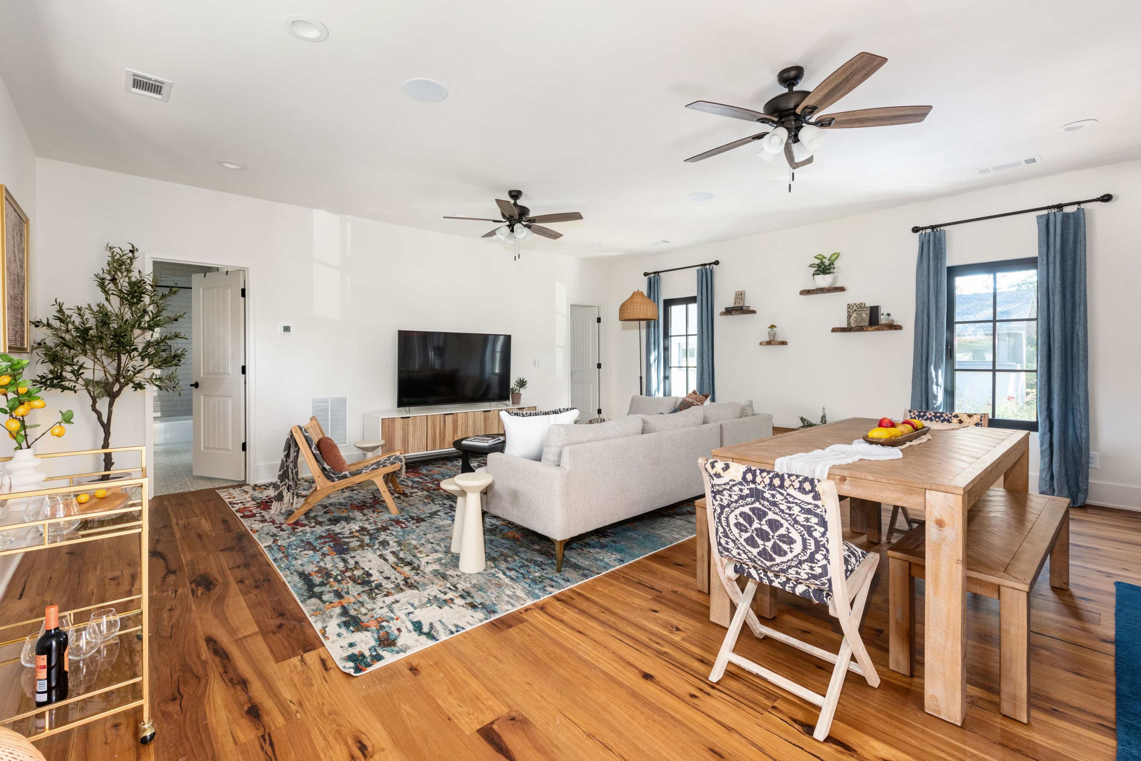 The living space features a light-filled room with a gray couch, wooden dining table, and a large television against a white wall.