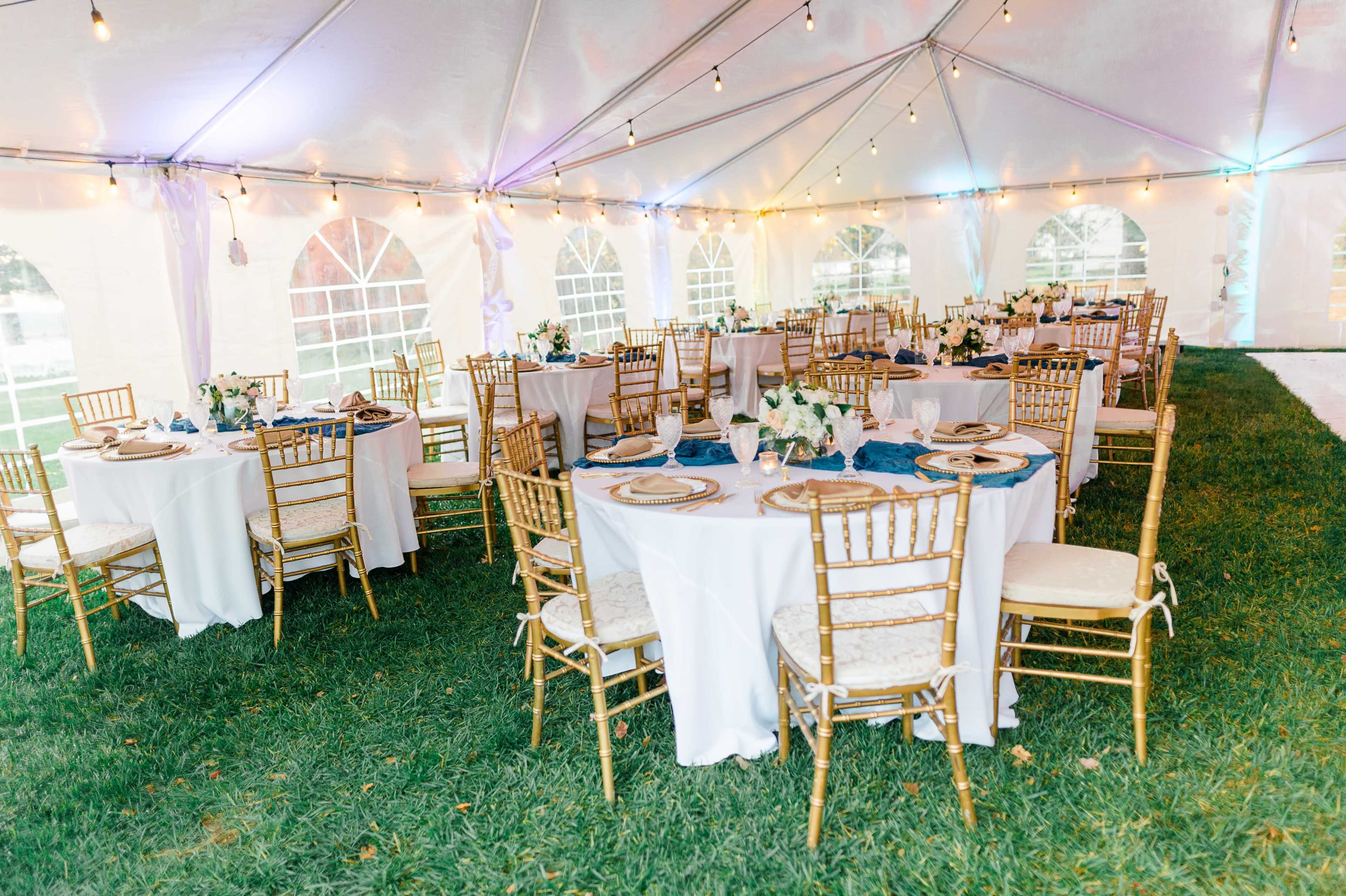 The image shows a tented outdoor dining area with round tables set for an event, adorned with white linens, gold chairs, and floral centerpieces.