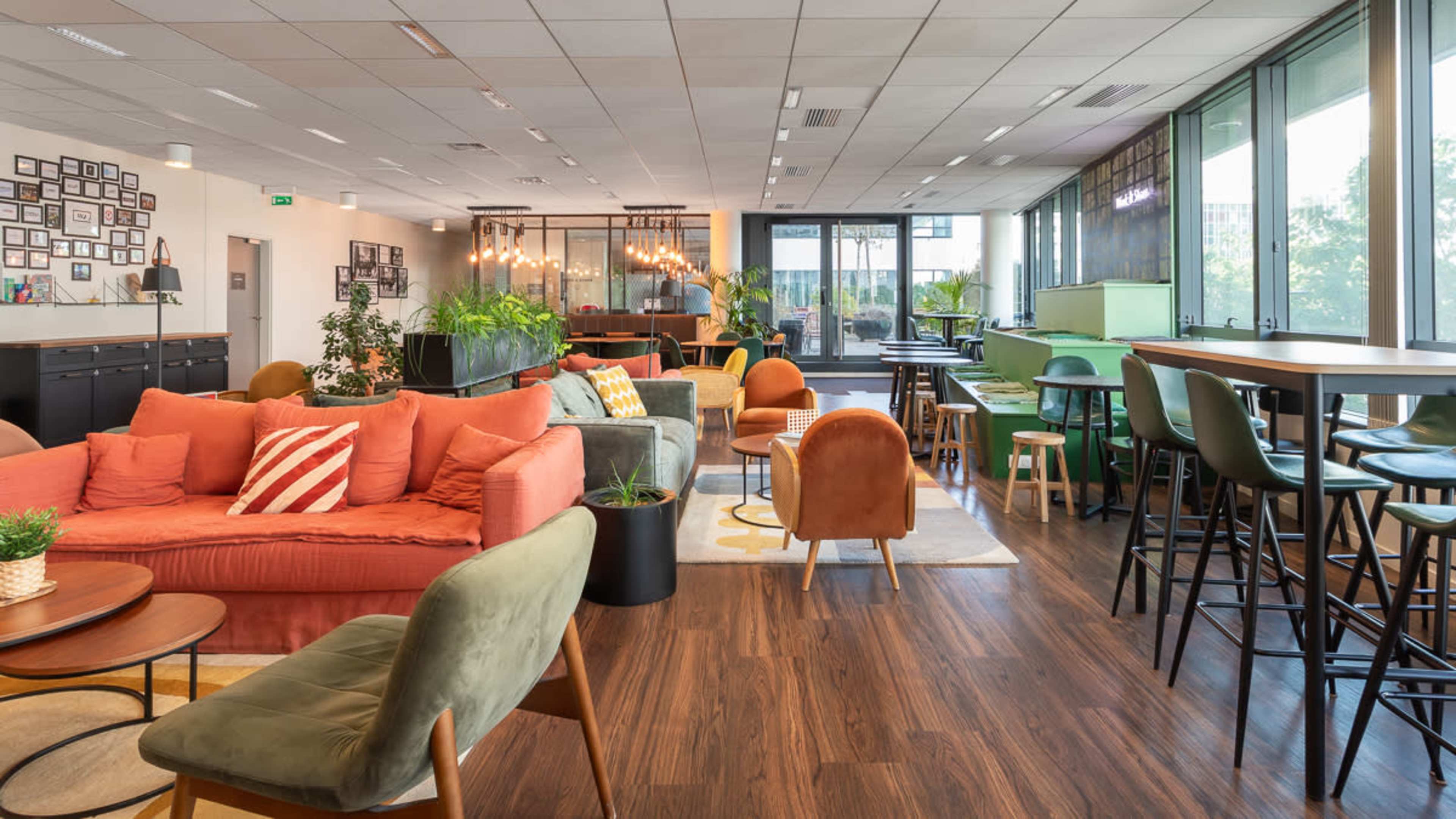 The image features a modern lounge area with a mix of seating options, including sofas and chairs, surrounded by large windows and indoor plants.