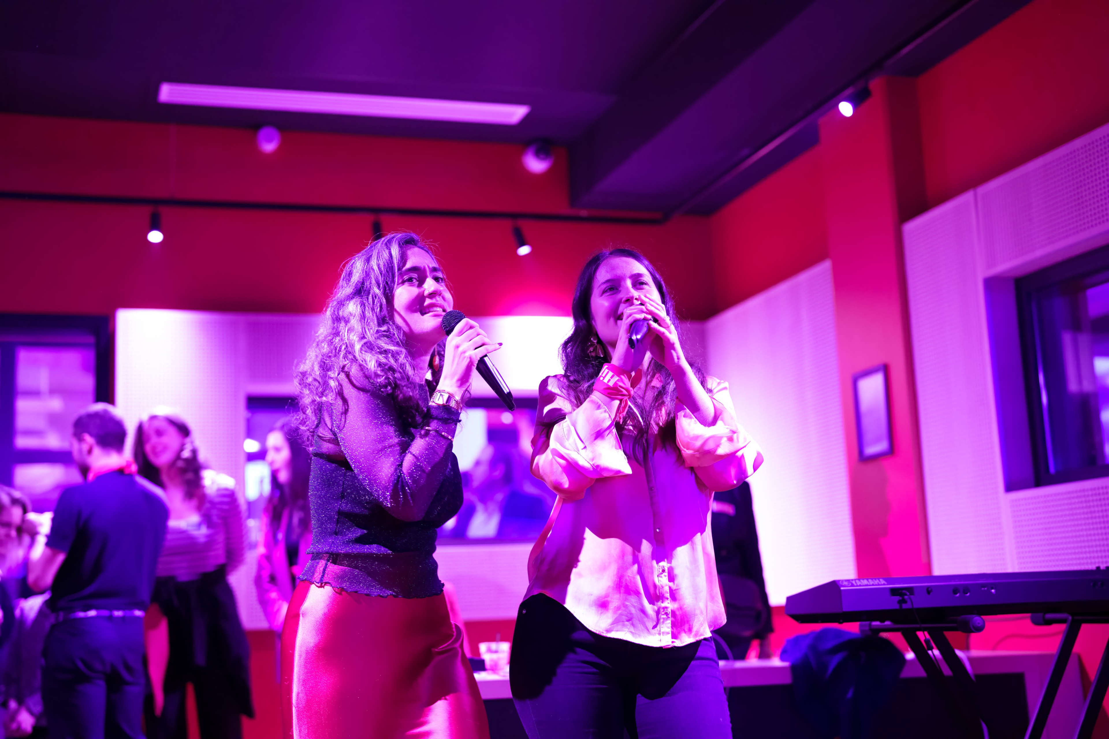 Two women sing into microphones on a stage while a crowd is gathered in the background, set in a brightly lit room with purple accents.