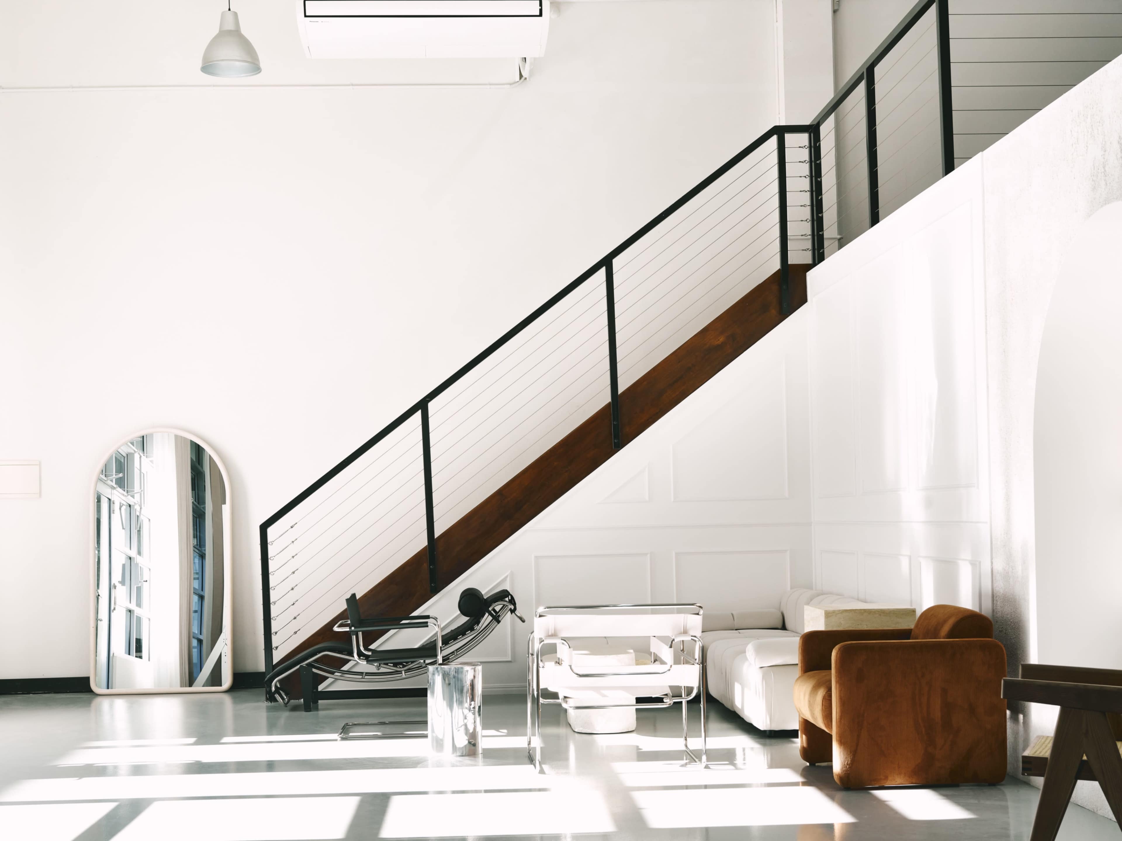 The image shows a bright, modern interior space with a staircase, a mirror, a workout machine, and a sofa.