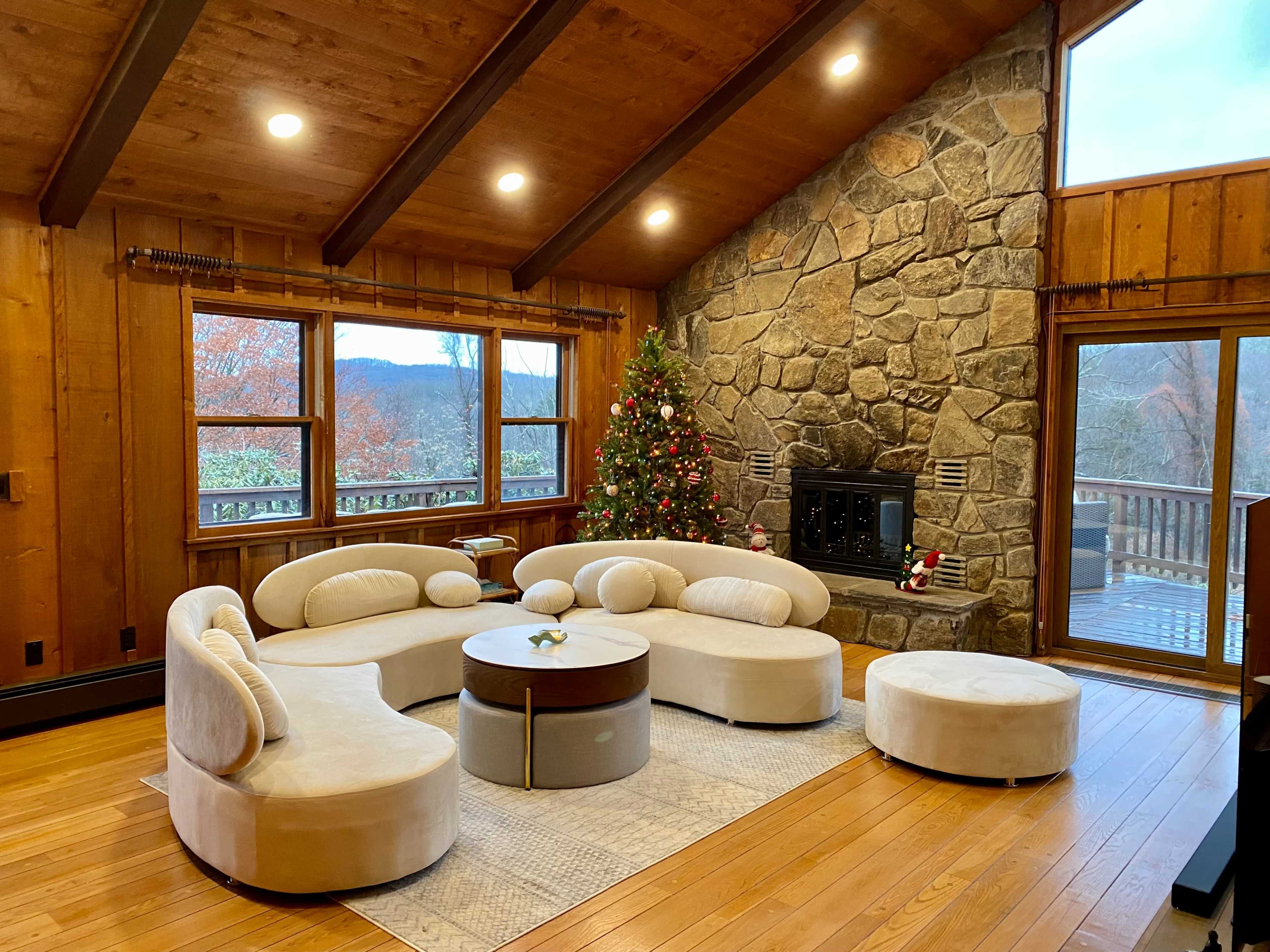 A cozy living room with a stone fireplace, a circular seating arrangement, and a decorated Christmas tree near large windows overlooking a scenic view.