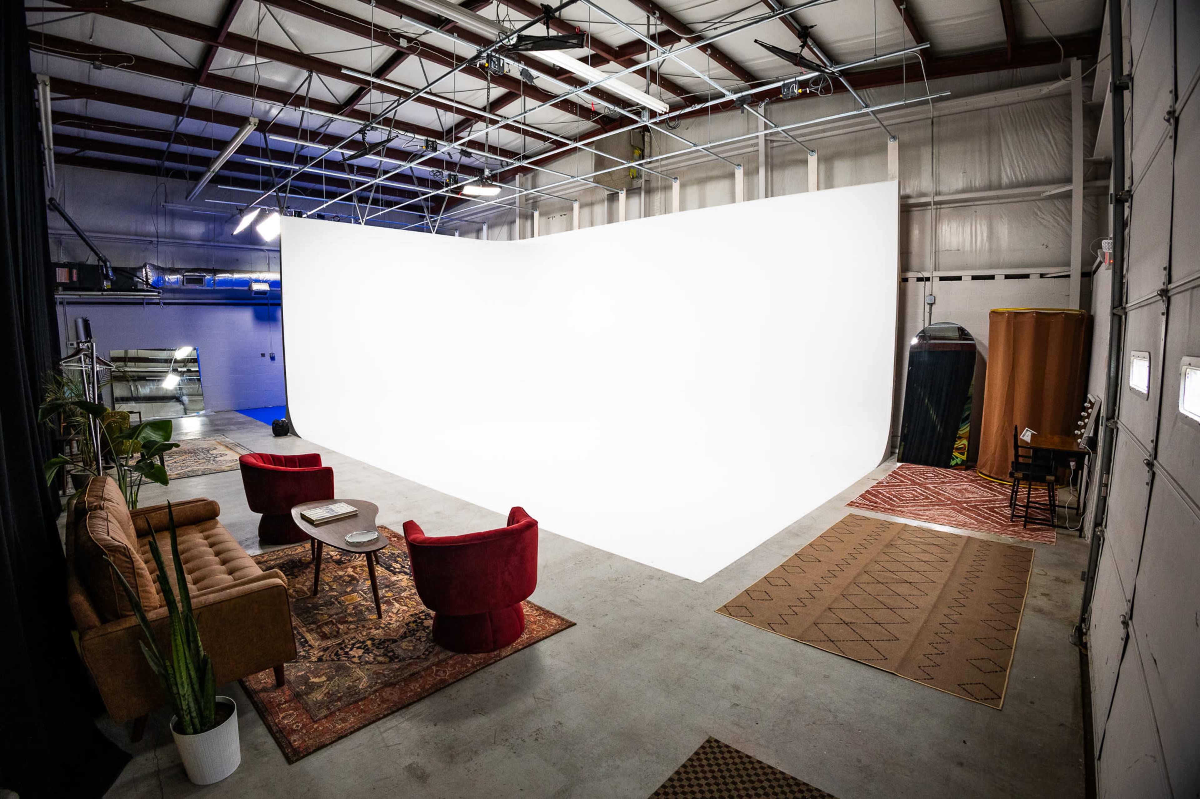 The image shows a spacious studio interior with a white backdrop, two seating areas featuring a brown couch and red chairs, and decorative rugs on the floor.