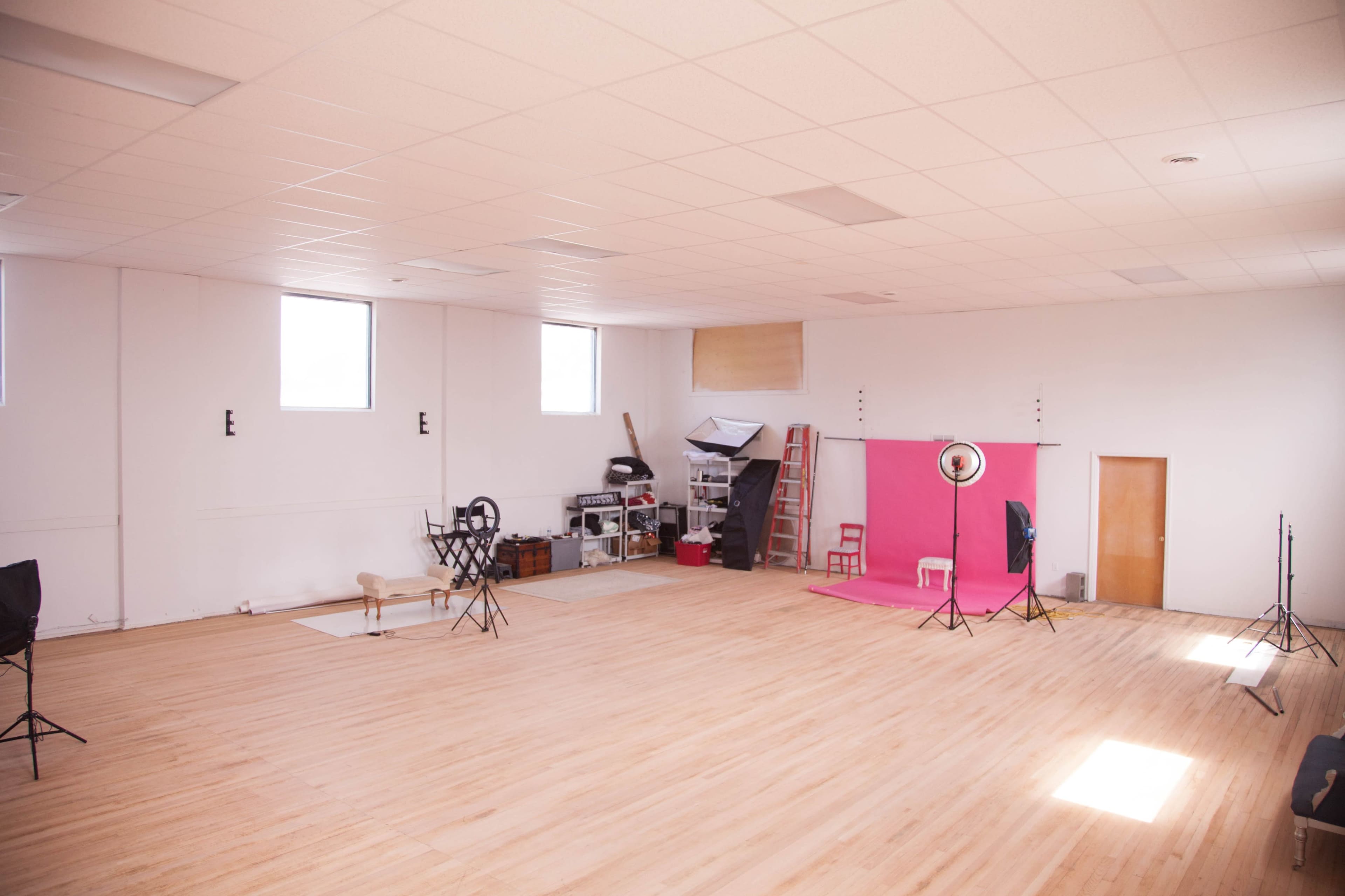 A spacious photography studio features hardwood floors, large windows, and a pink backdrop setup with lighting equipment on one side.