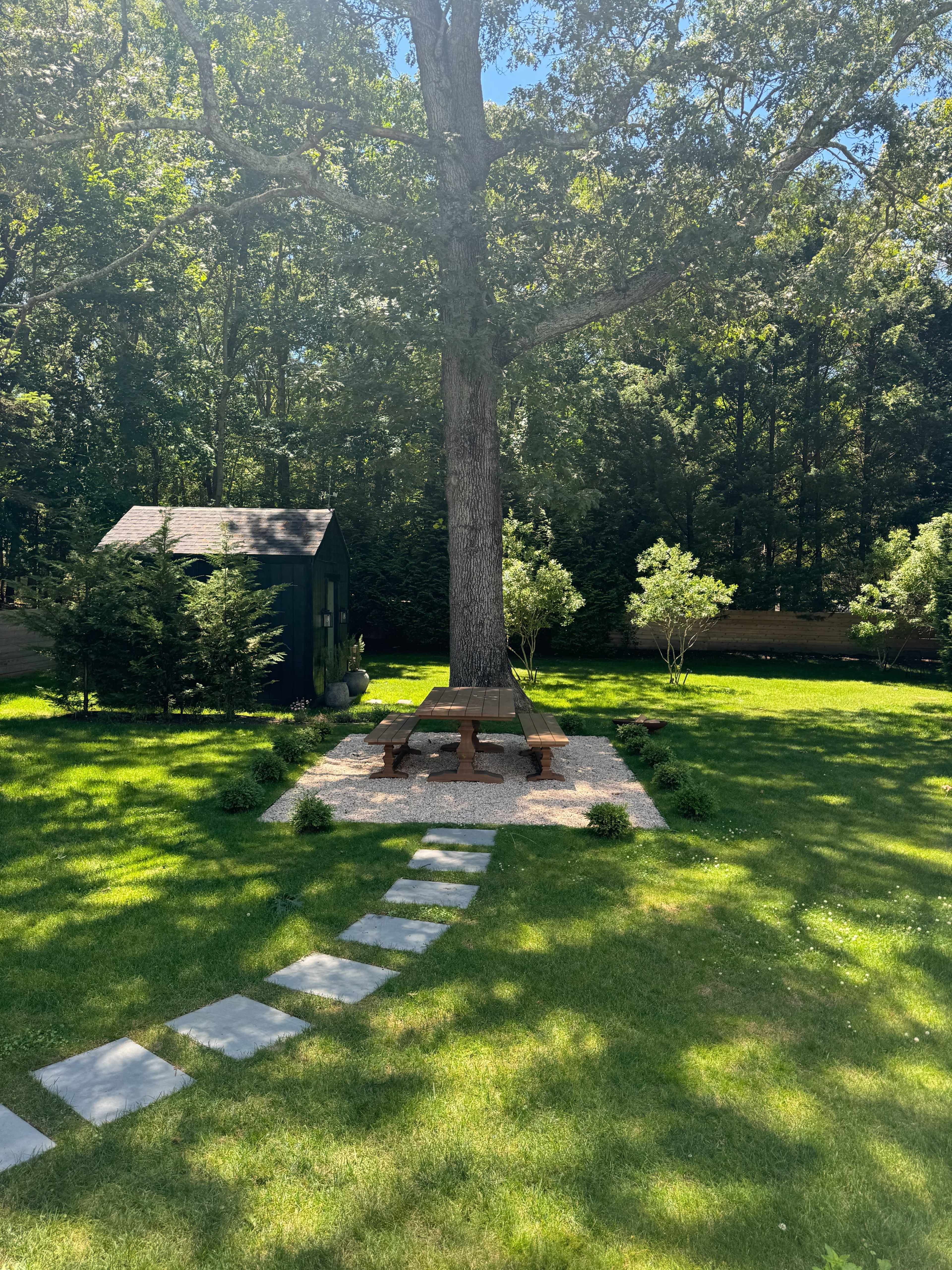 A round picnic table is situated under a large tree in a grassy yard, with a stone pathway leading towards it and a small shed in the background.