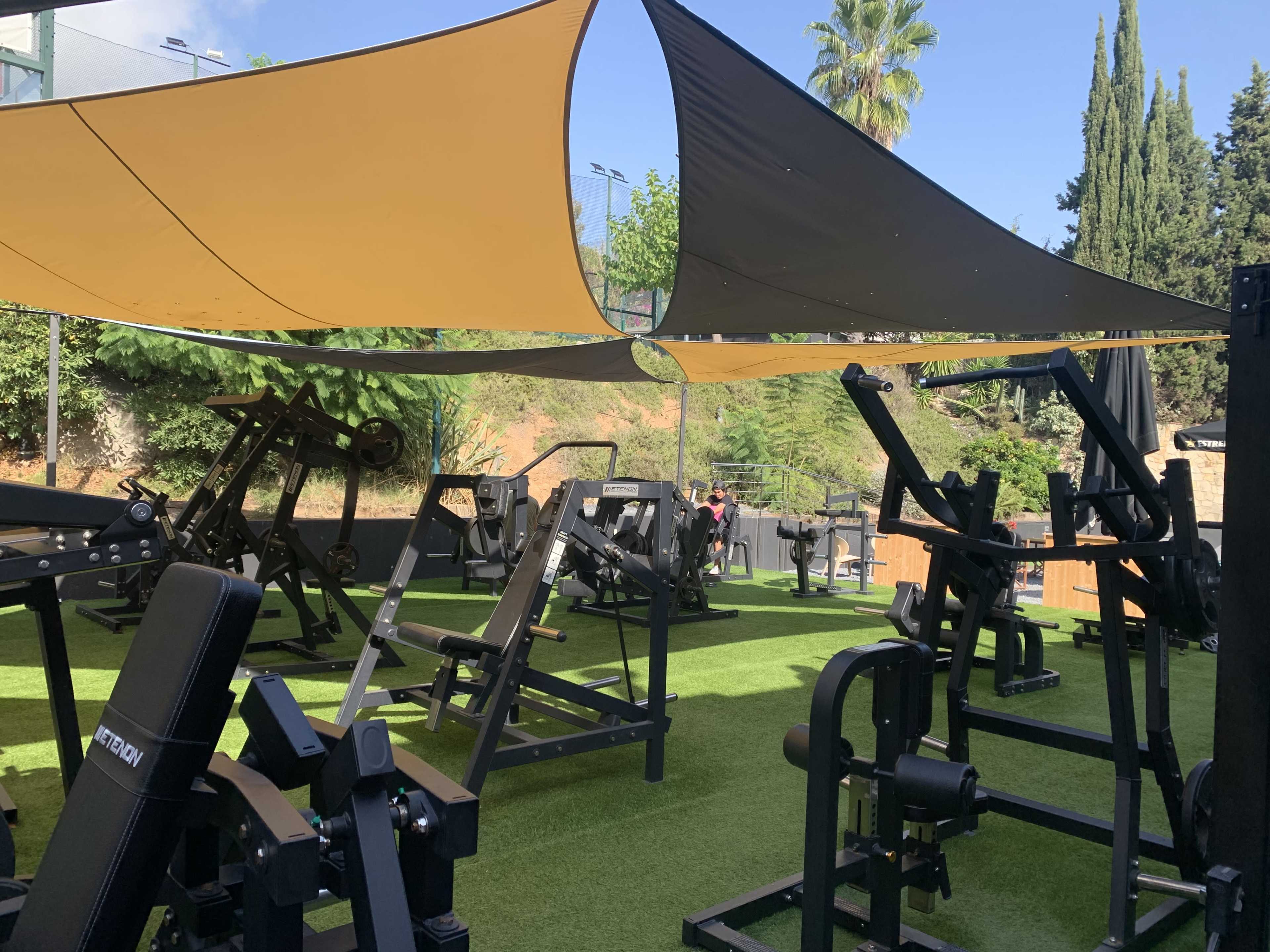 An outdoor gym area with various weightlifting equipment under shaded canopies and surrounded by greenery.