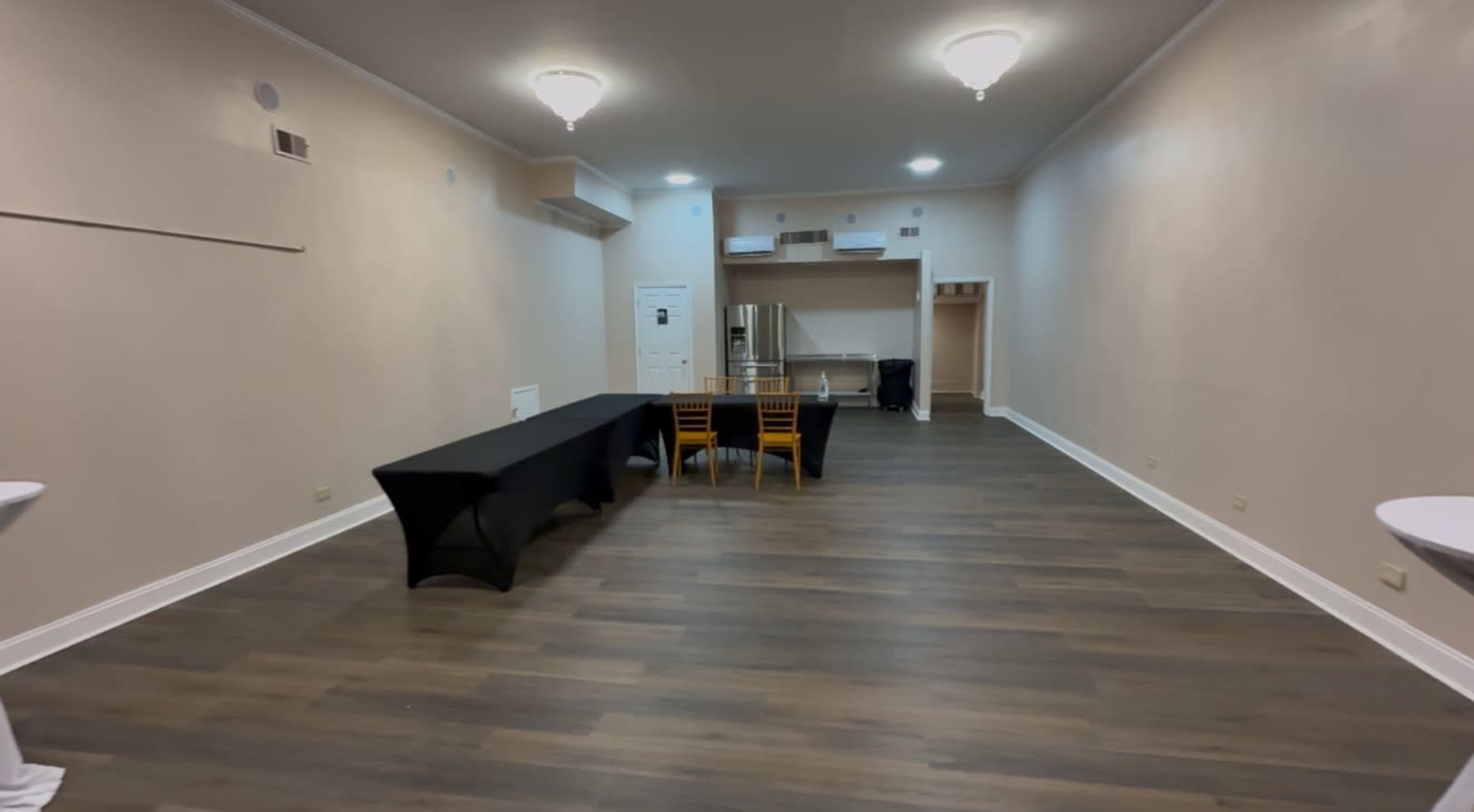 The image shows a spacious, empty room with wooden flooring, a few tables and chairs arranged in the center, and a kitchenette area in the background.