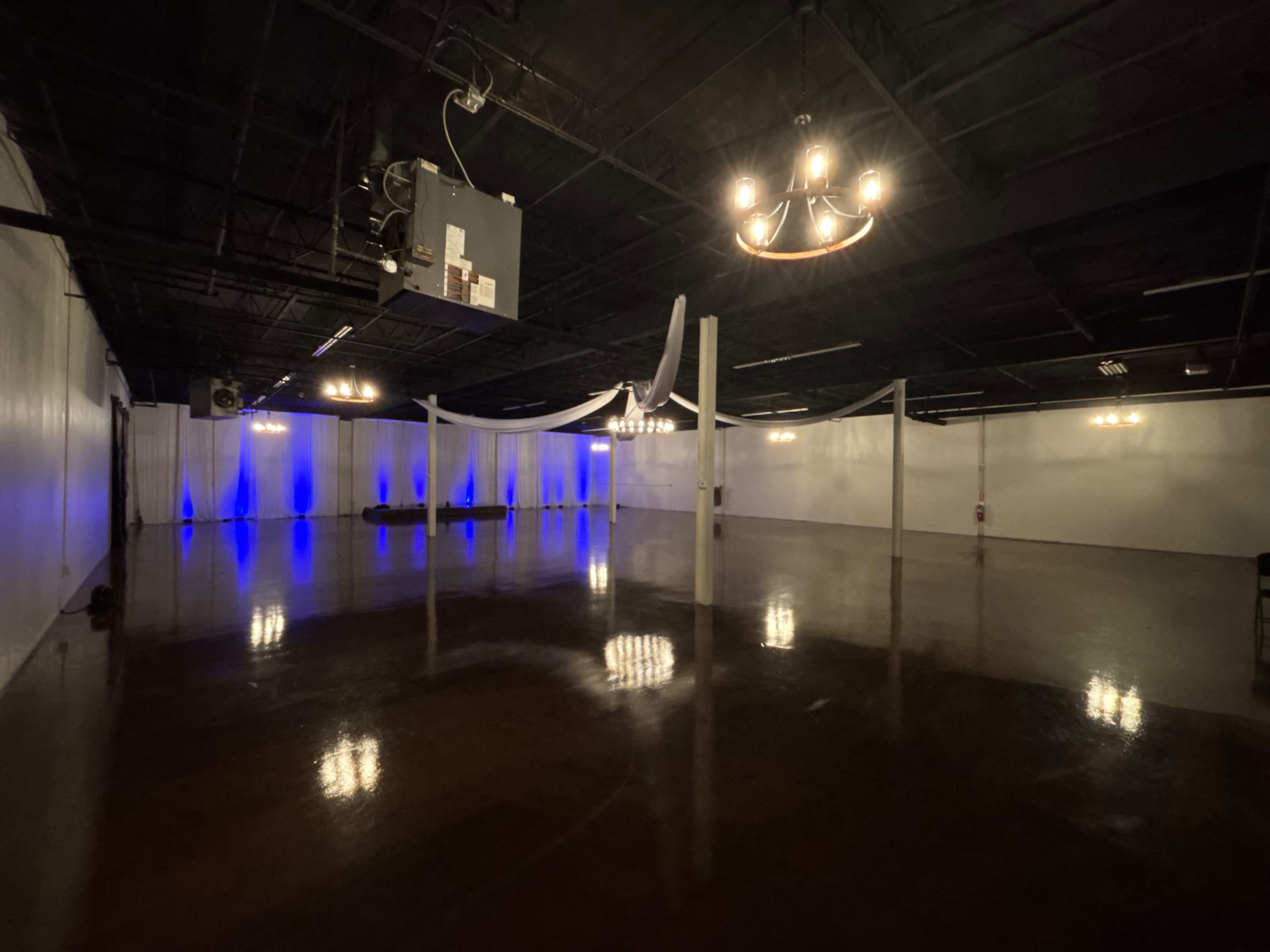 The image shows a spacious, well-lit event hall with a polished floor, white walls, and decorative lighting, including chandeliers and blue accents.