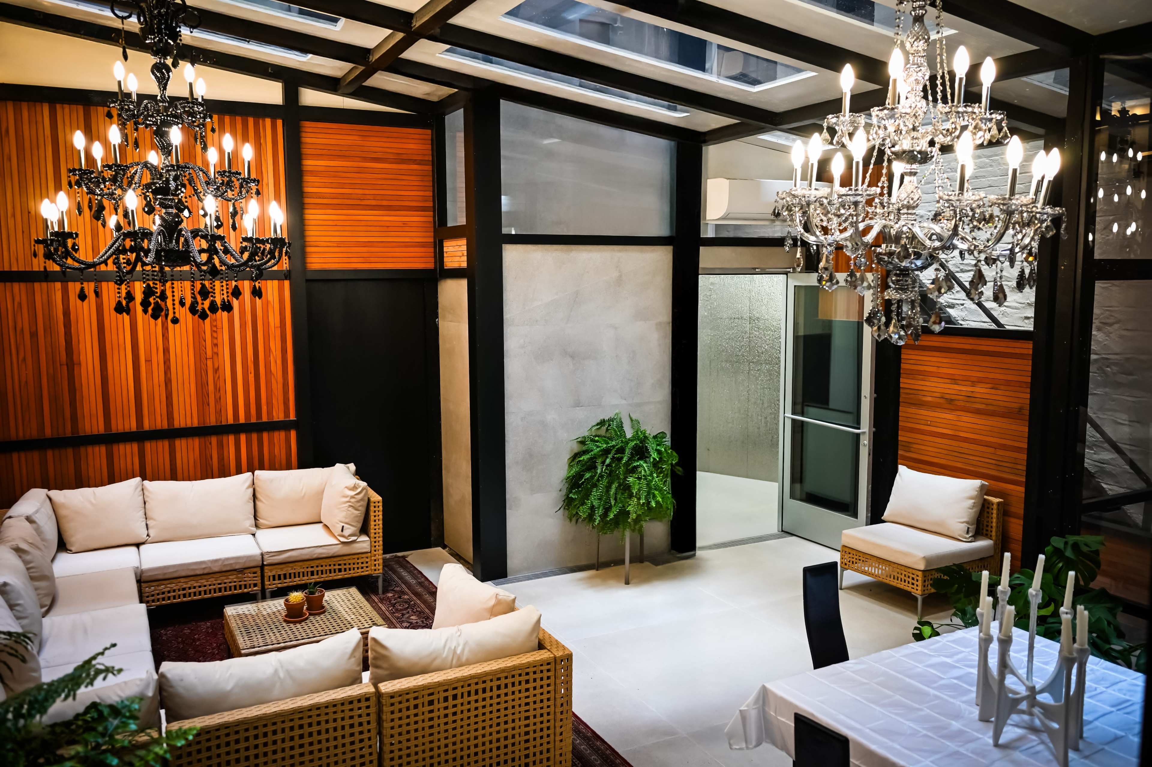 The image shows a modern indoor lounge area featuring wicker seating, chandeliers, and a glass ceiling with wooden paneling.
