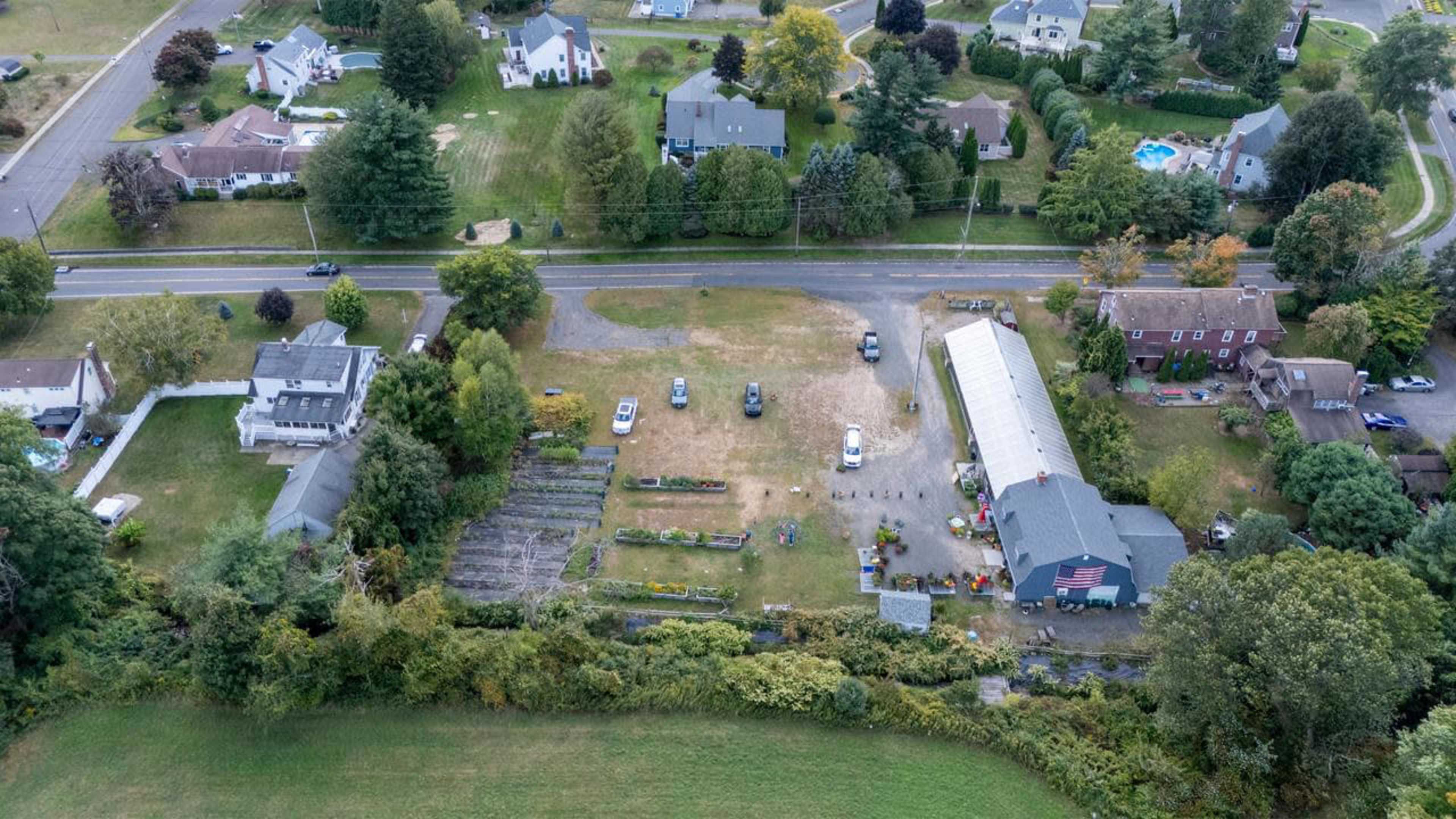 The aerial image shows a rural neighborhood with houses, gardens, and a parking area, bordered by a road and surrounded by trees.
