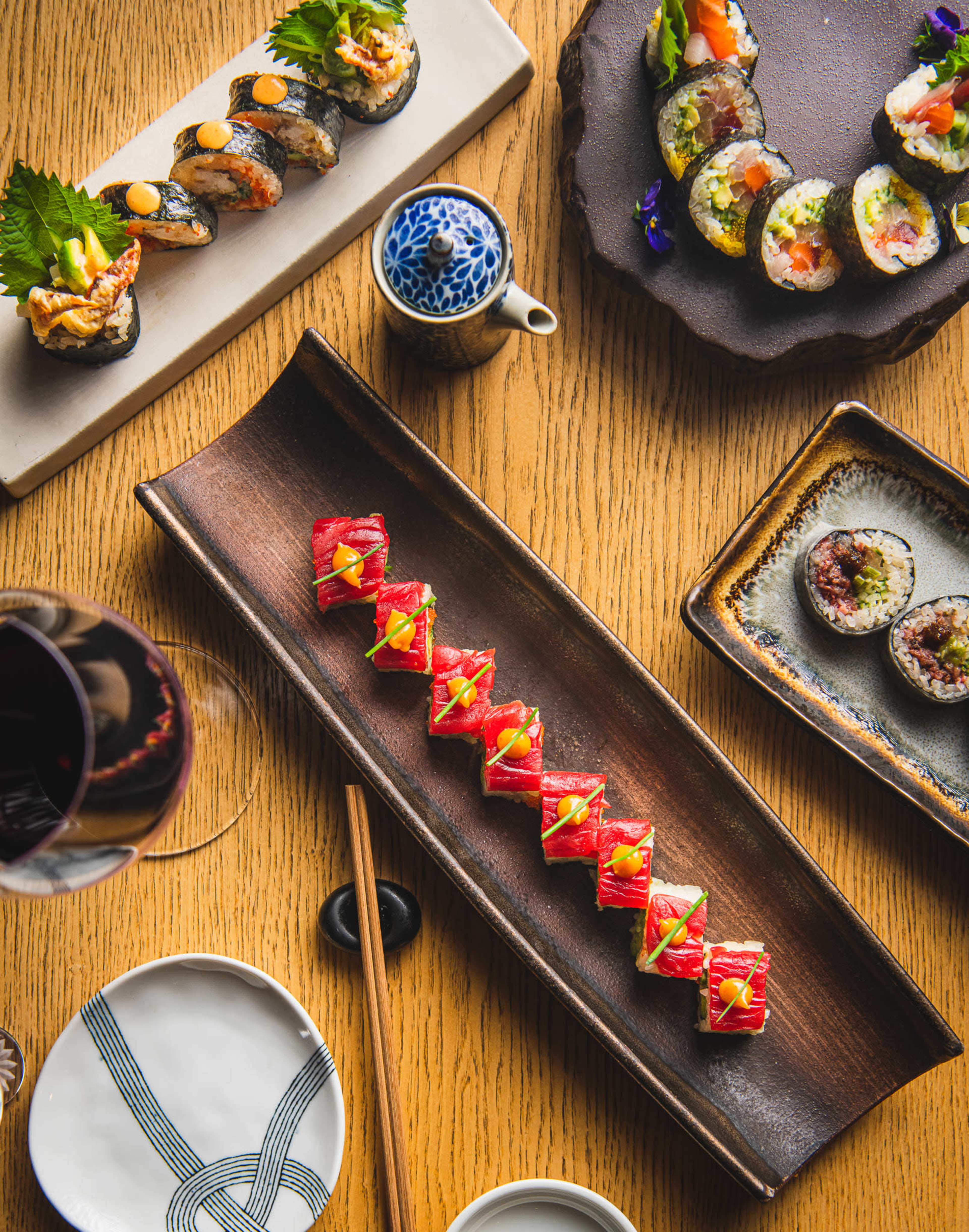 The image shows an assortment of sushi dishes, including rolls and sashimi, arranged on wooden and ceramic plates with chopsticks and a small teapot in the background.