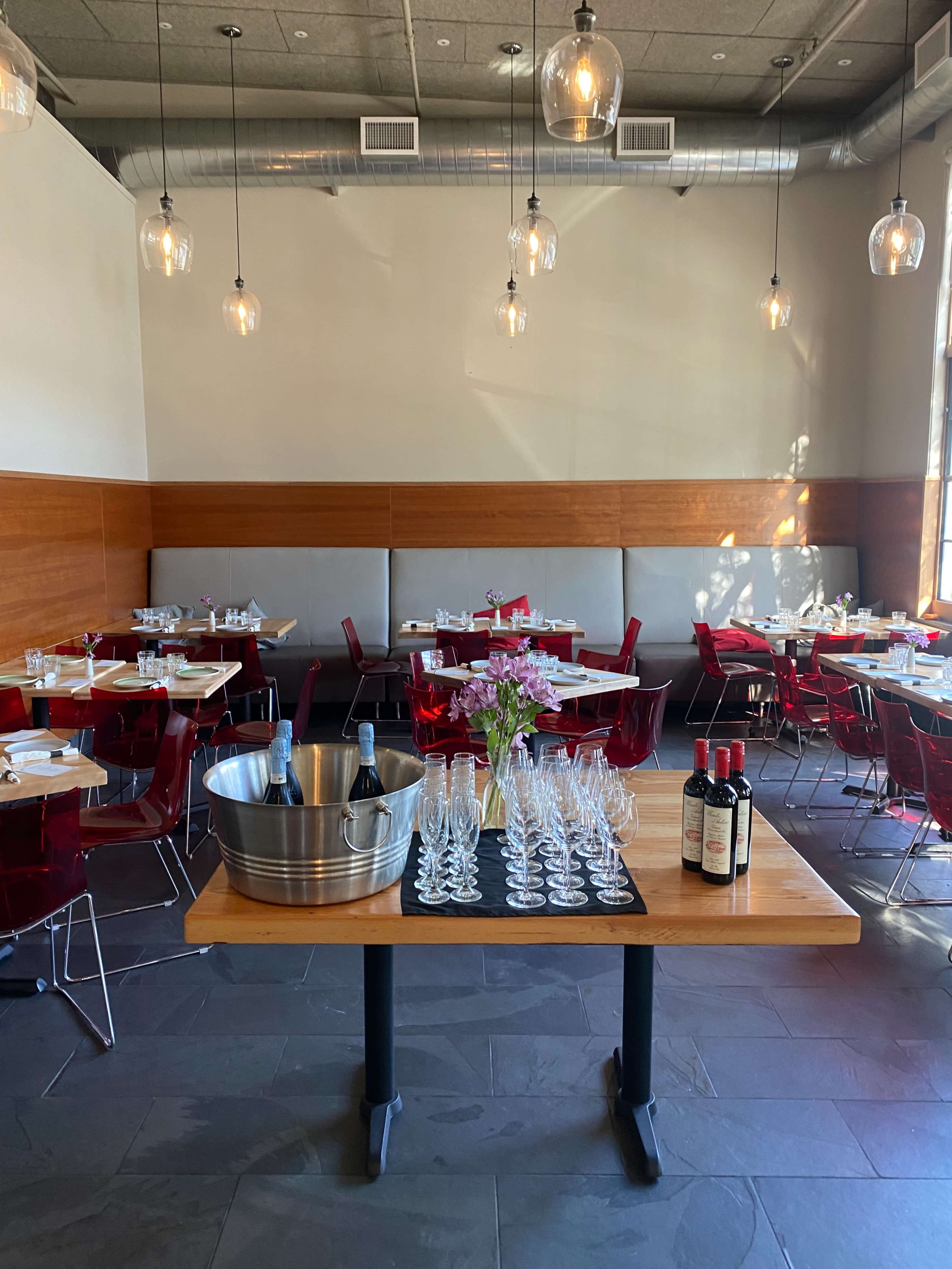 A dining area features empty tables with red chairs, a centerpiece table with a bucket of champagne, and a display of glasses and wine bottles.