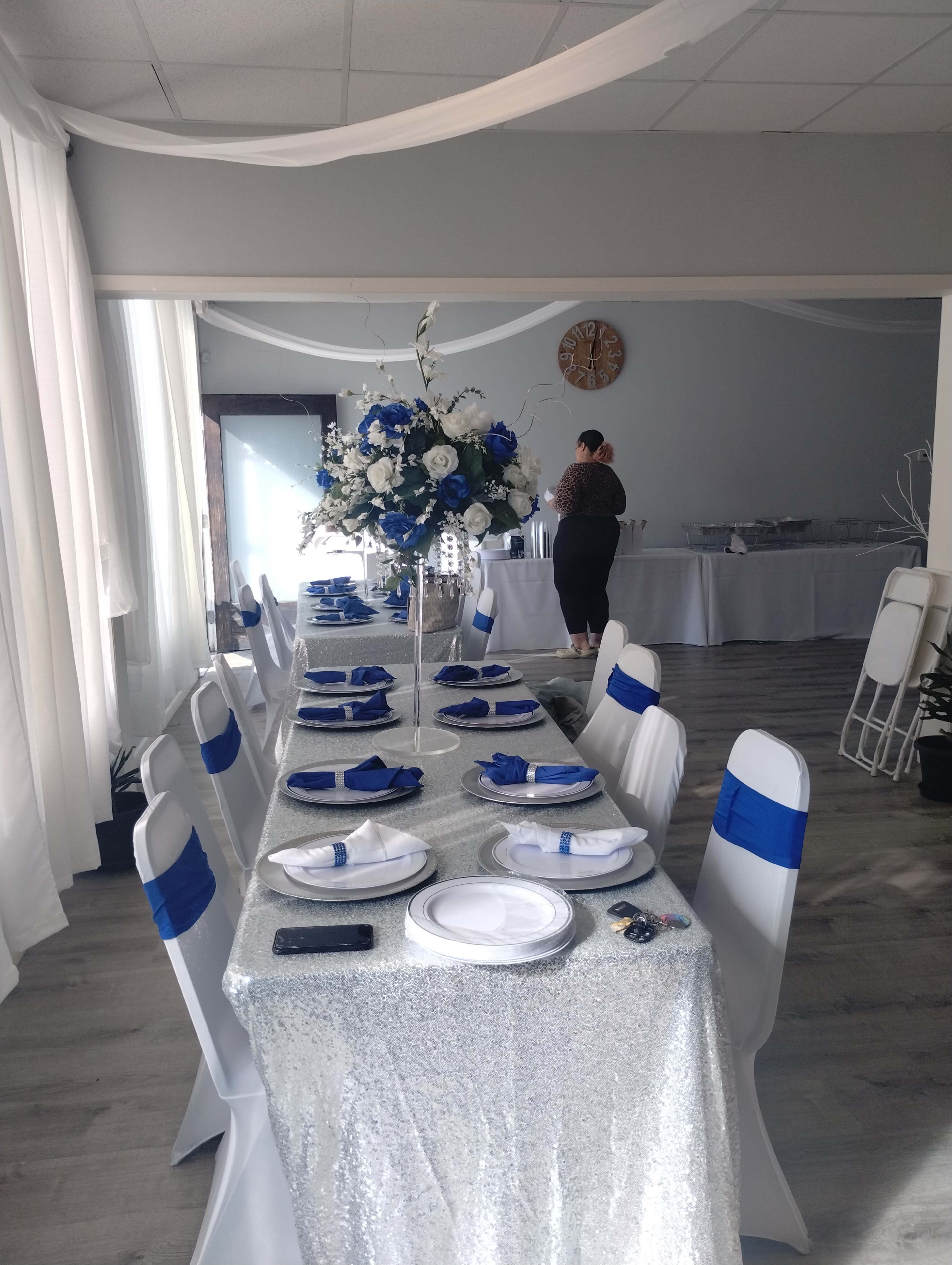 A long banquet table adorned with a sparkly silver tablecloth and blue decorations is set up in a brightly lit event space, while a person is seen arranging items in the background.