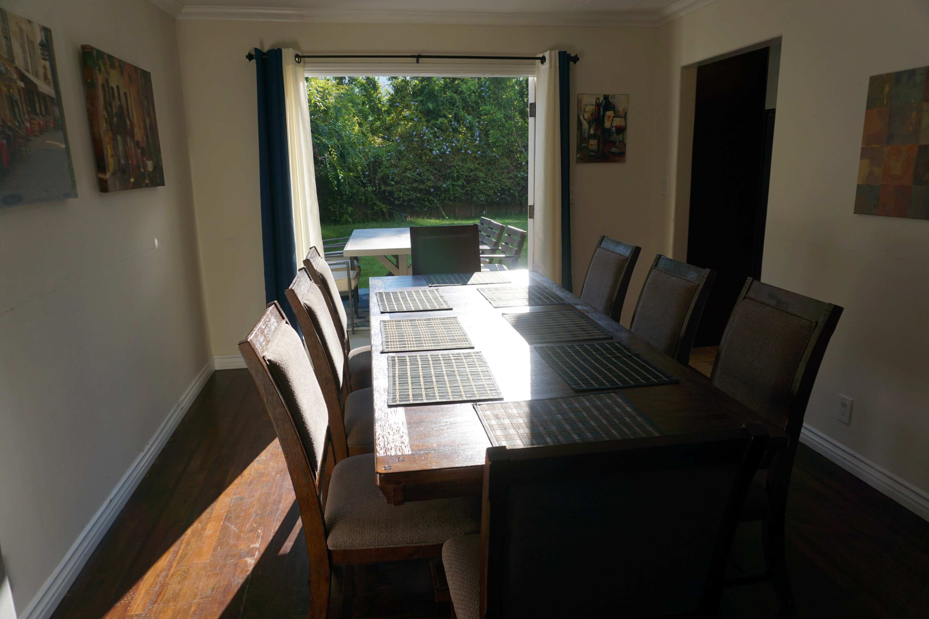 A wooden dining table with eight chairs is set against a window that overlooks a green backyard.