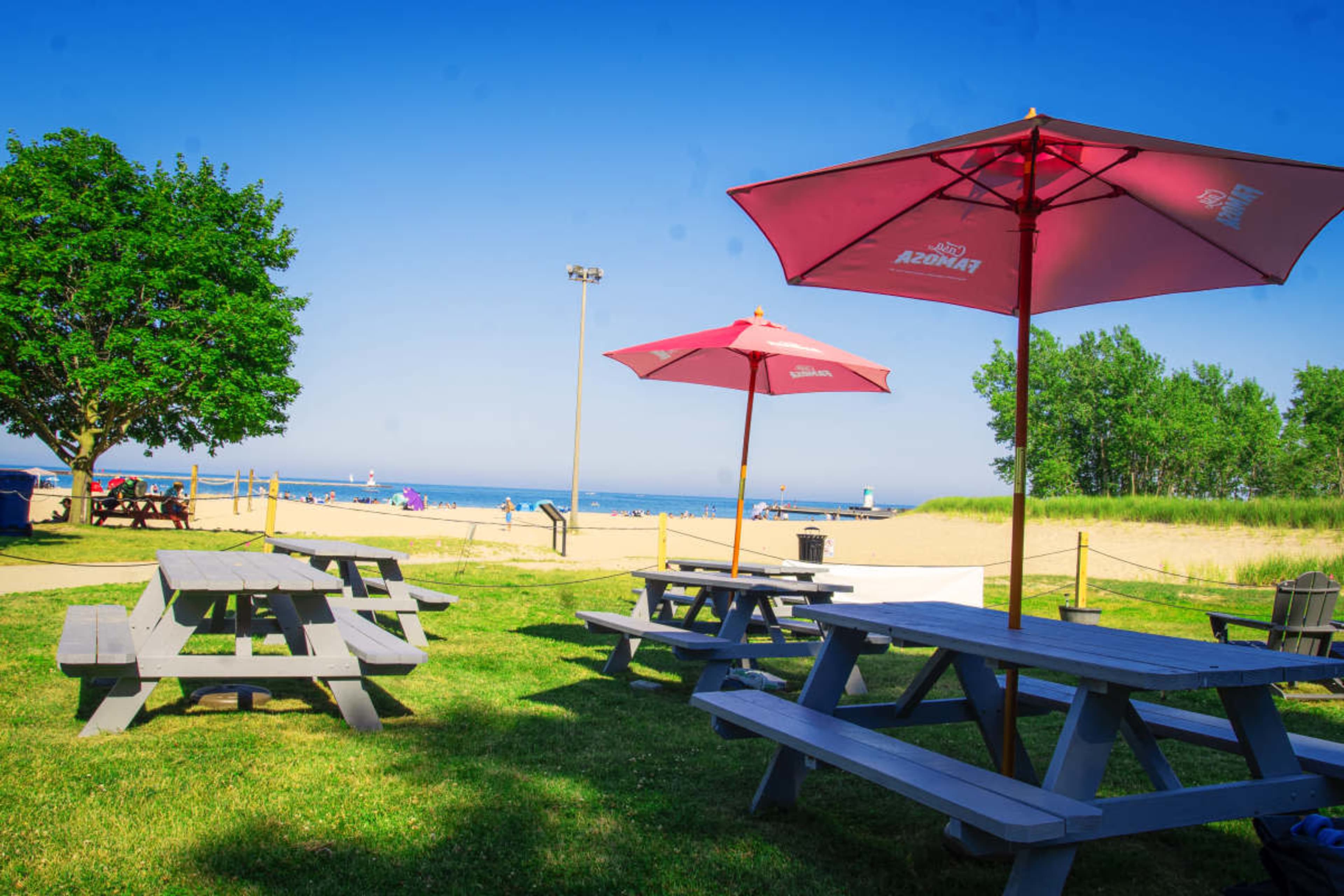 A park area features gray picnic tables and red umbrellas with a beach and ocean in the background.