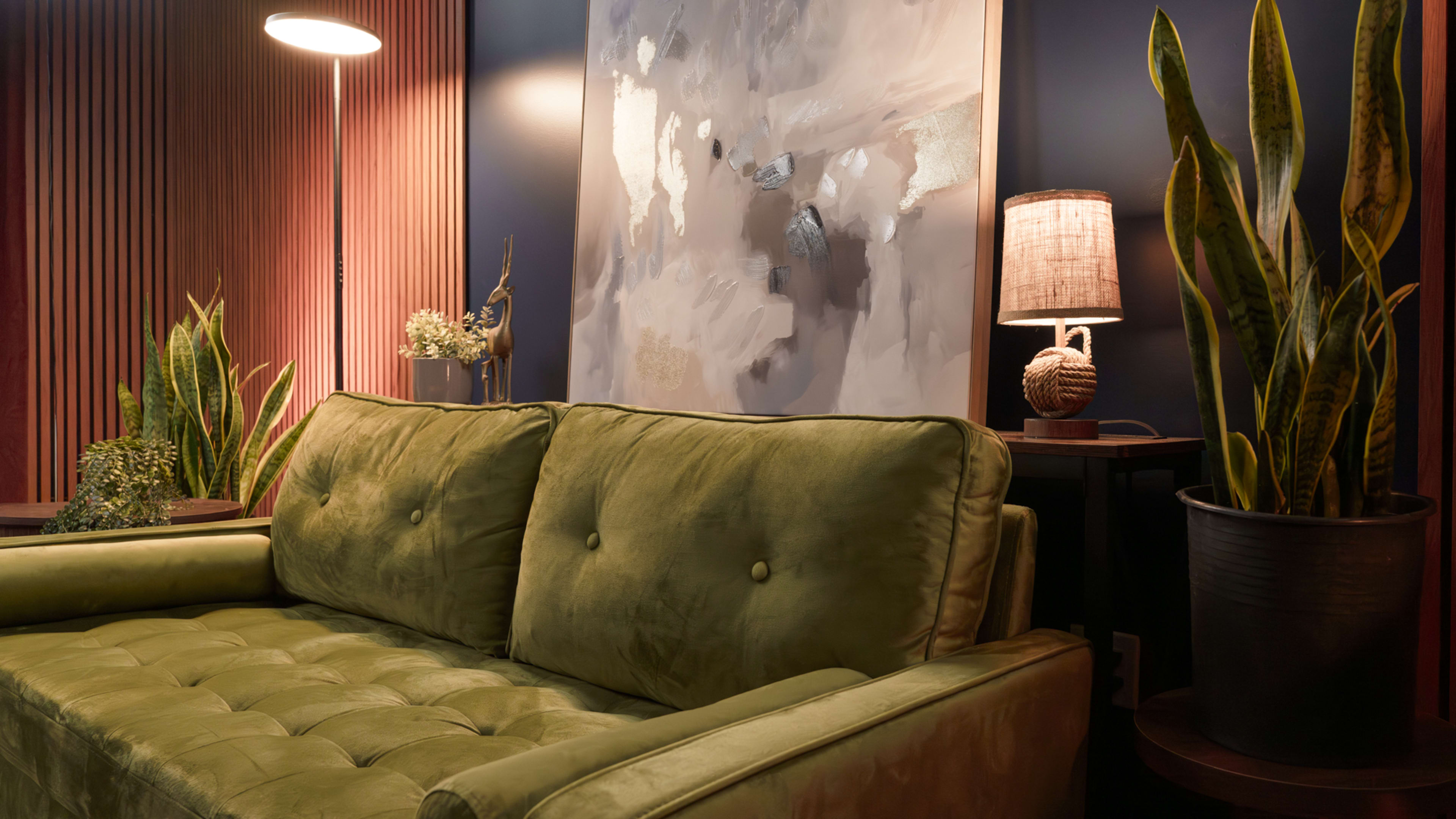 The image shows a cozy living area featuring a green velvet sofa, a large abstract painting on the wall, tall plants, and lamps providing warm lighting.