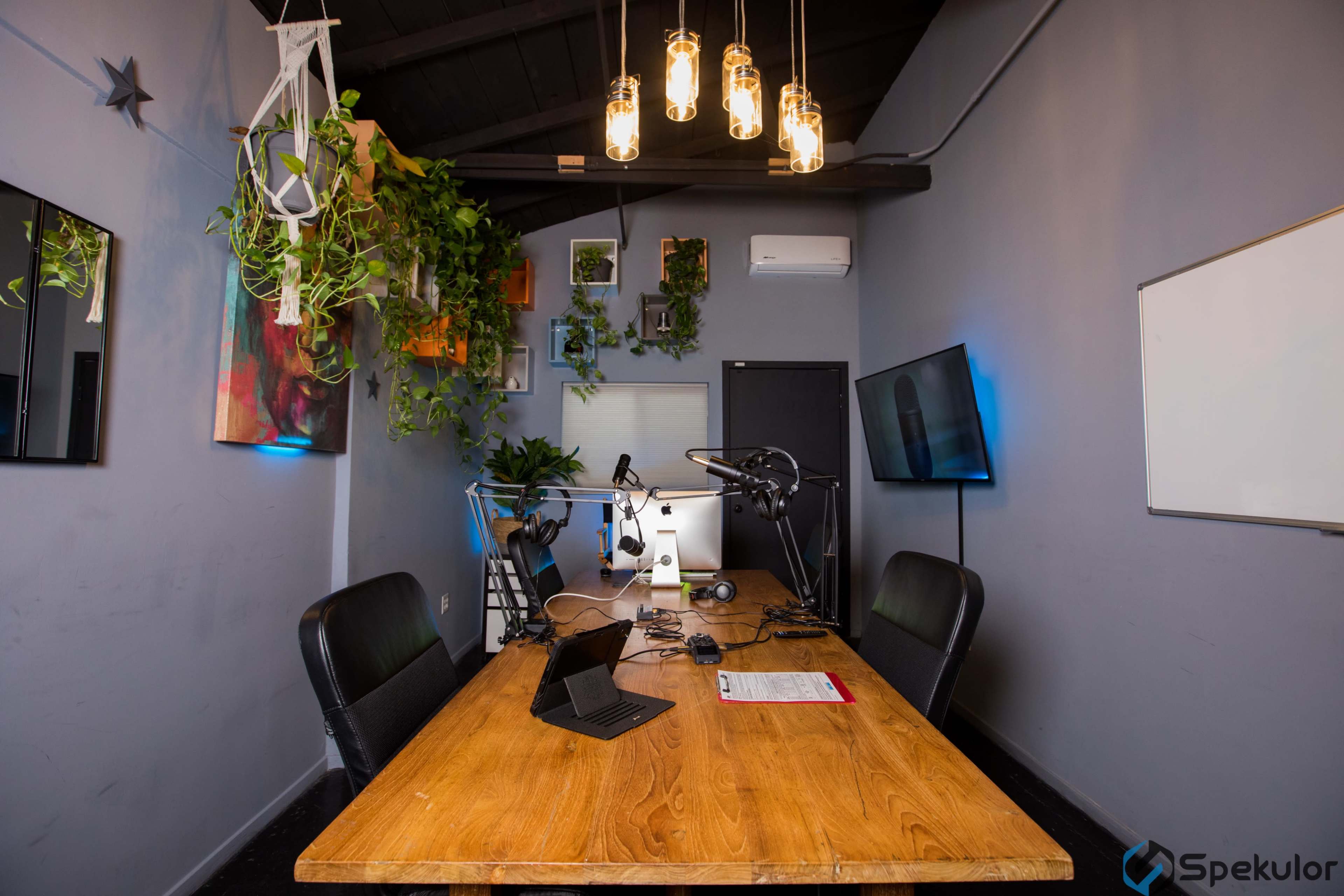 A modern office space features a wooden table with microphones and multiple monitors, surrounded by green plants and decorative wall art.
