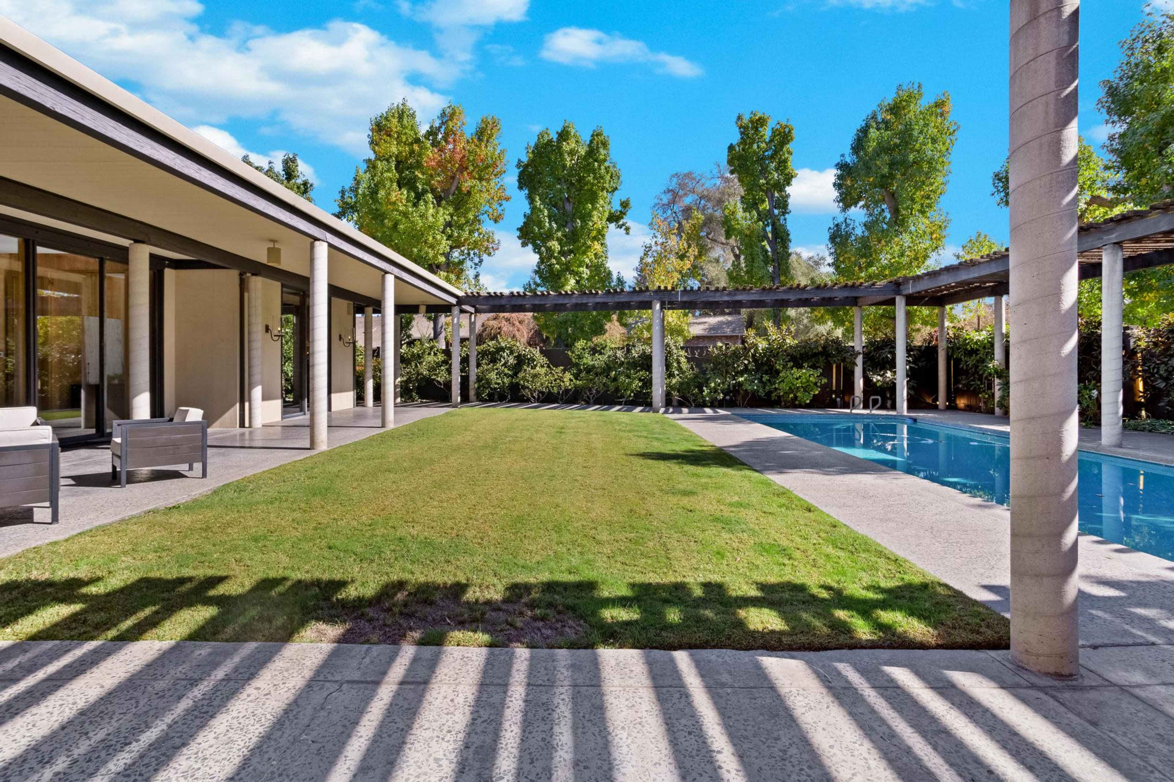 A landscaped backyard with a pool, green grass, and trees, framed by a modern house with a covered patio.