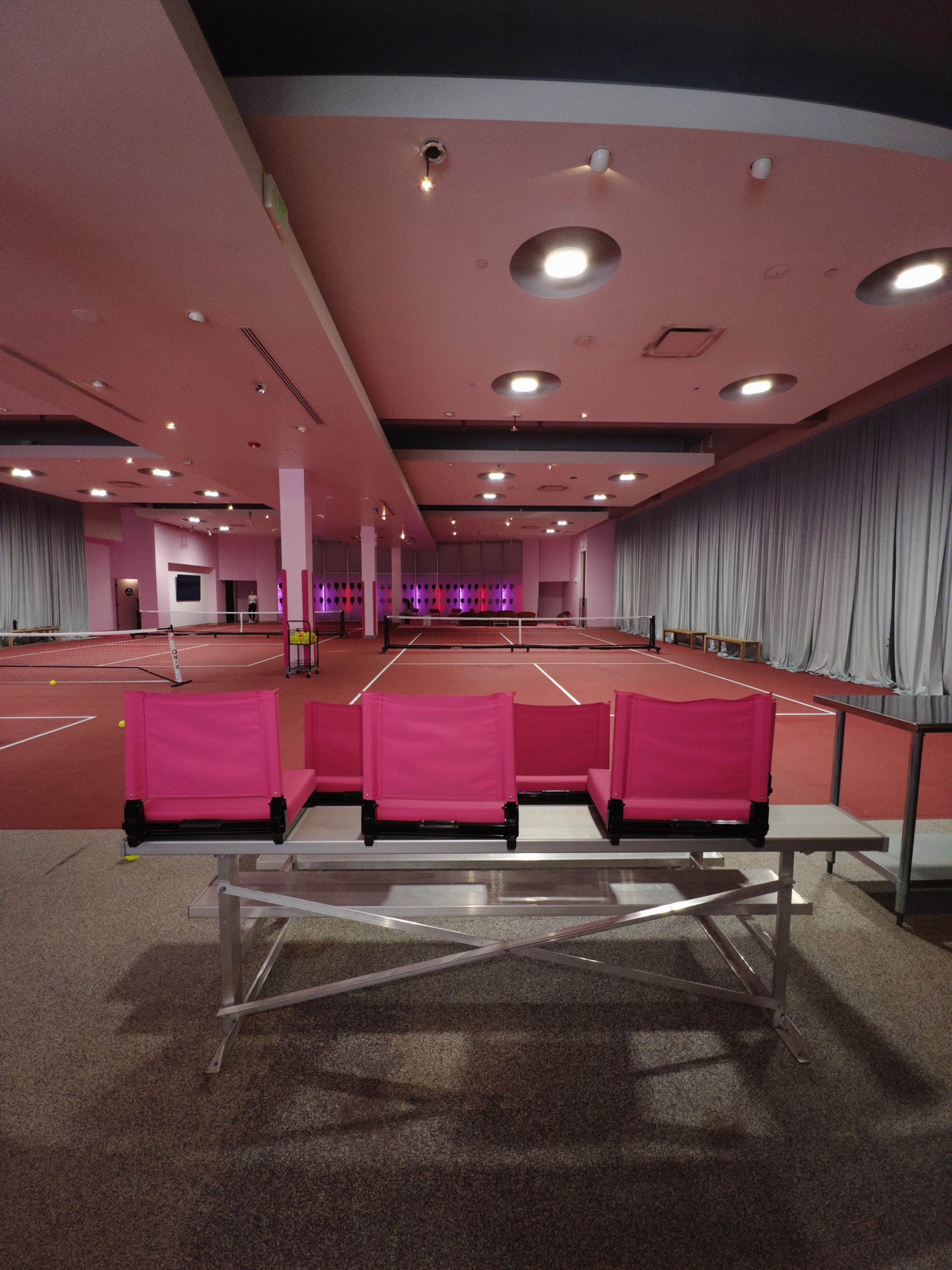 The image shows a spacious indoor tennis facility featuring several courts, with pink seating in the foreground and colorful lighting enhancing the atmosphere in the background.