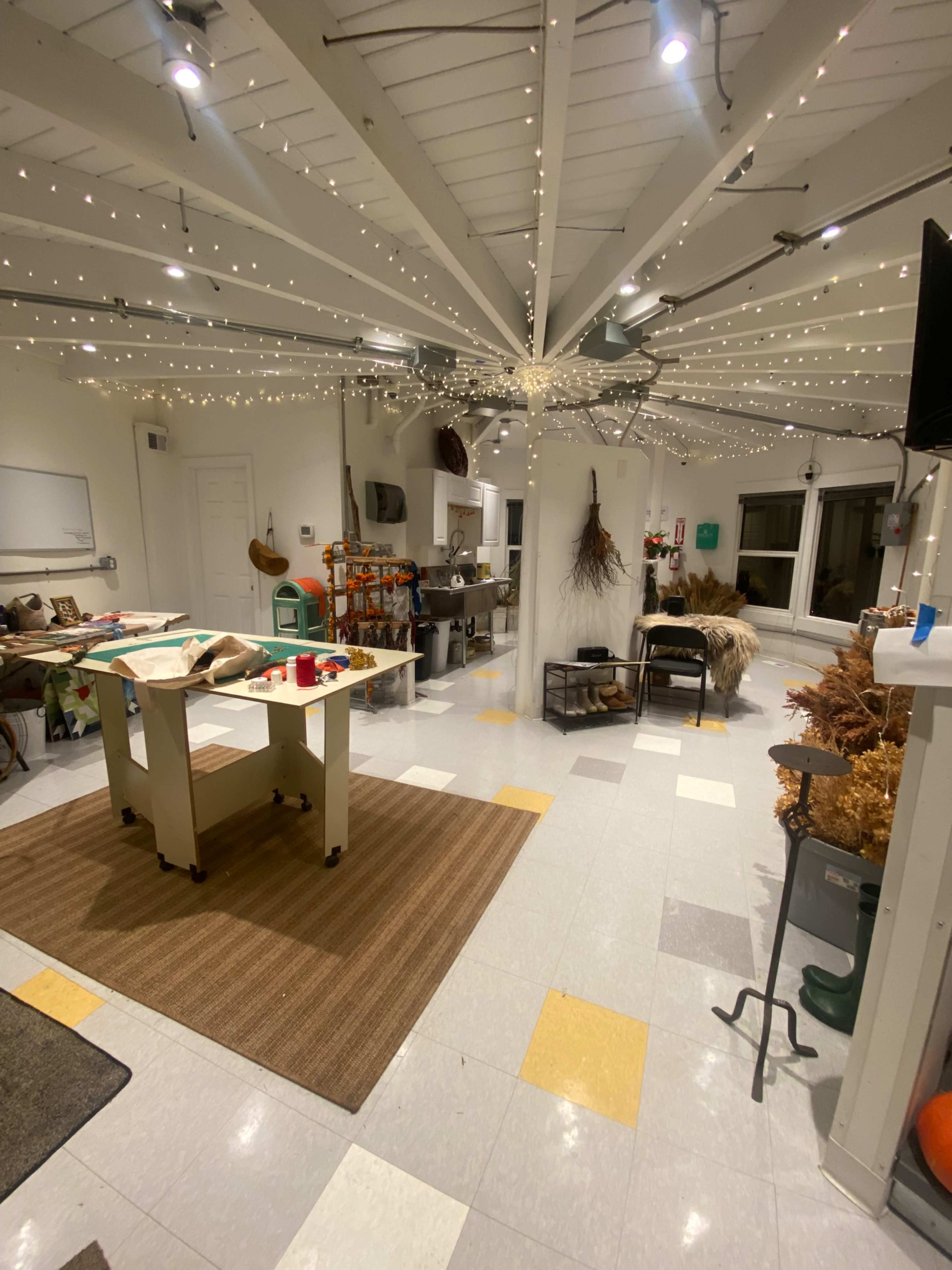 The image shows a spacious room with a kitchen area on the left, equipped with various appliances, a central table, and decorative lighting strung from the ceiling.