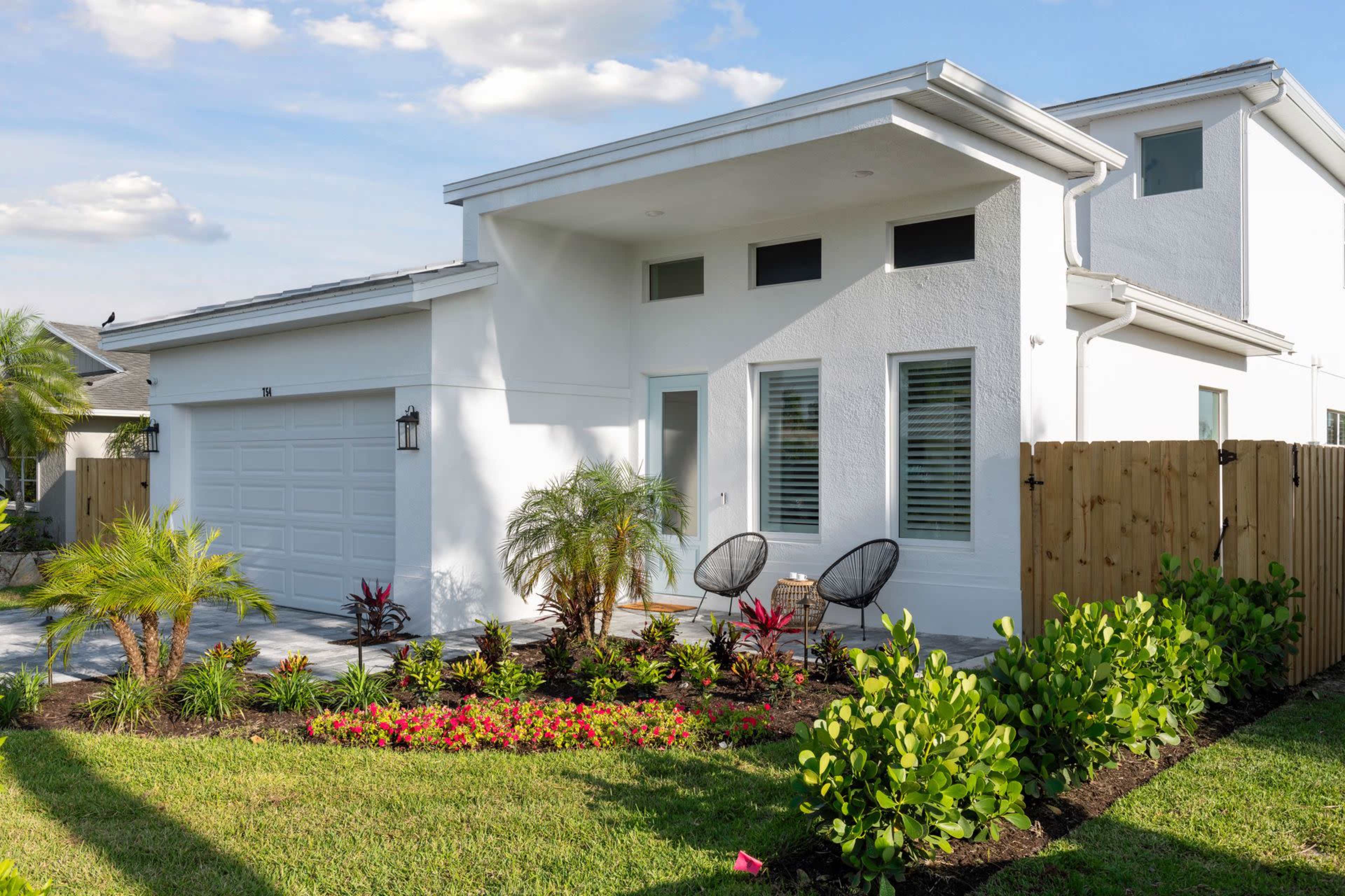 A modern white house with a landscaped front yard featuring colorful flowers and two chairs on the porch.