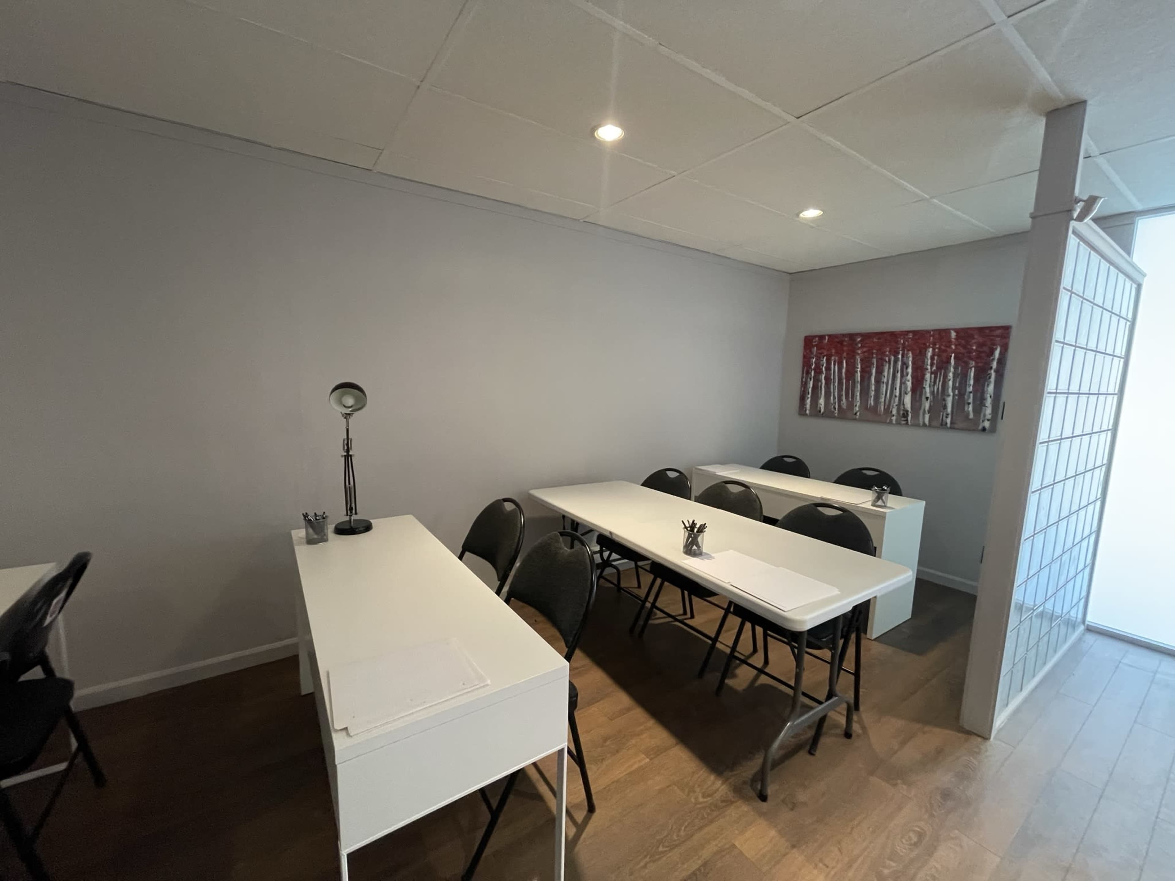 A small, well-lit classroom with two rows of white tables and black chairs, alongside a lamp and some artwork on the gray wall.