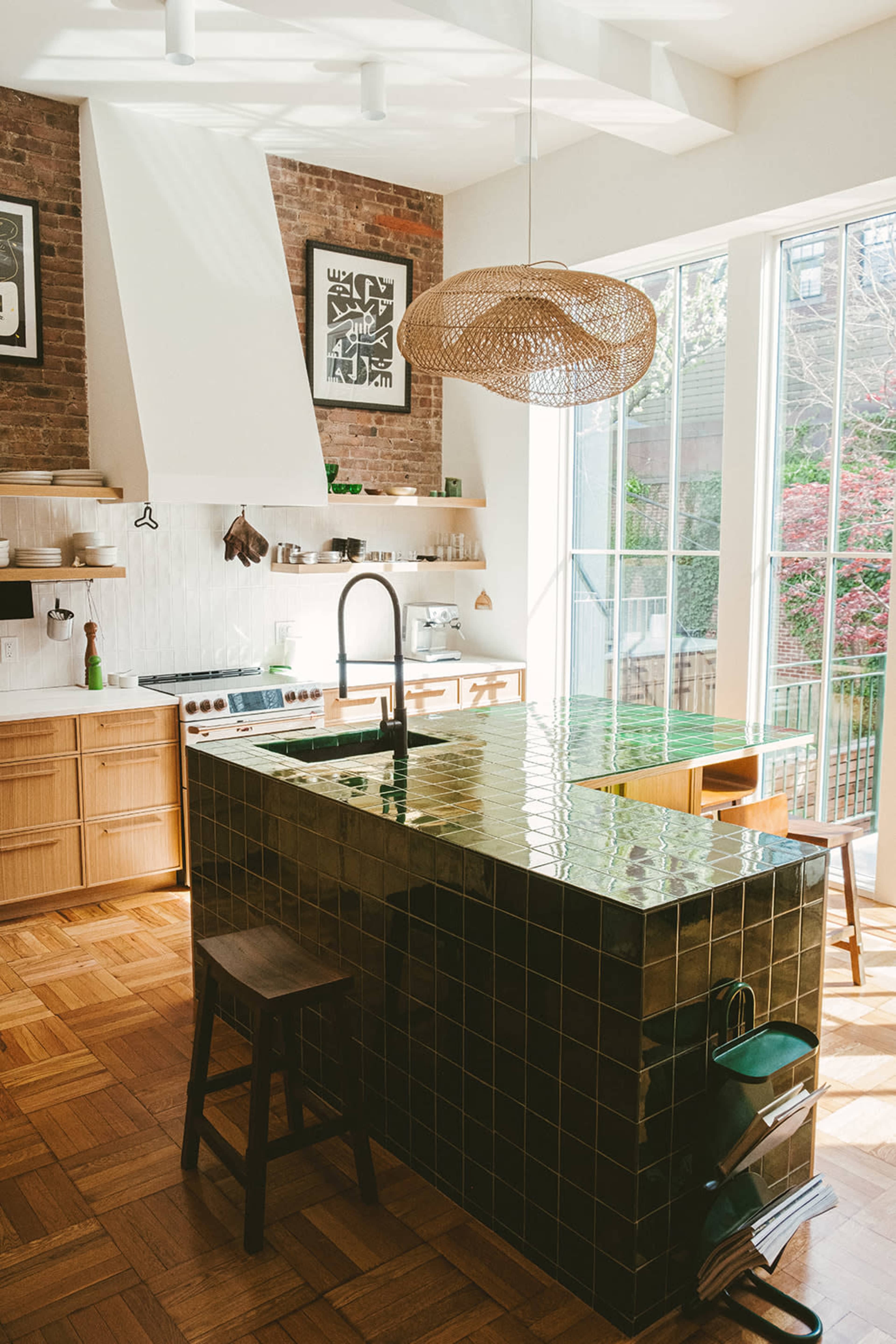 The image shows a modern kitchen with a green tiled island, wooden cabinetry, and large windows allowing natural light to flood the space.