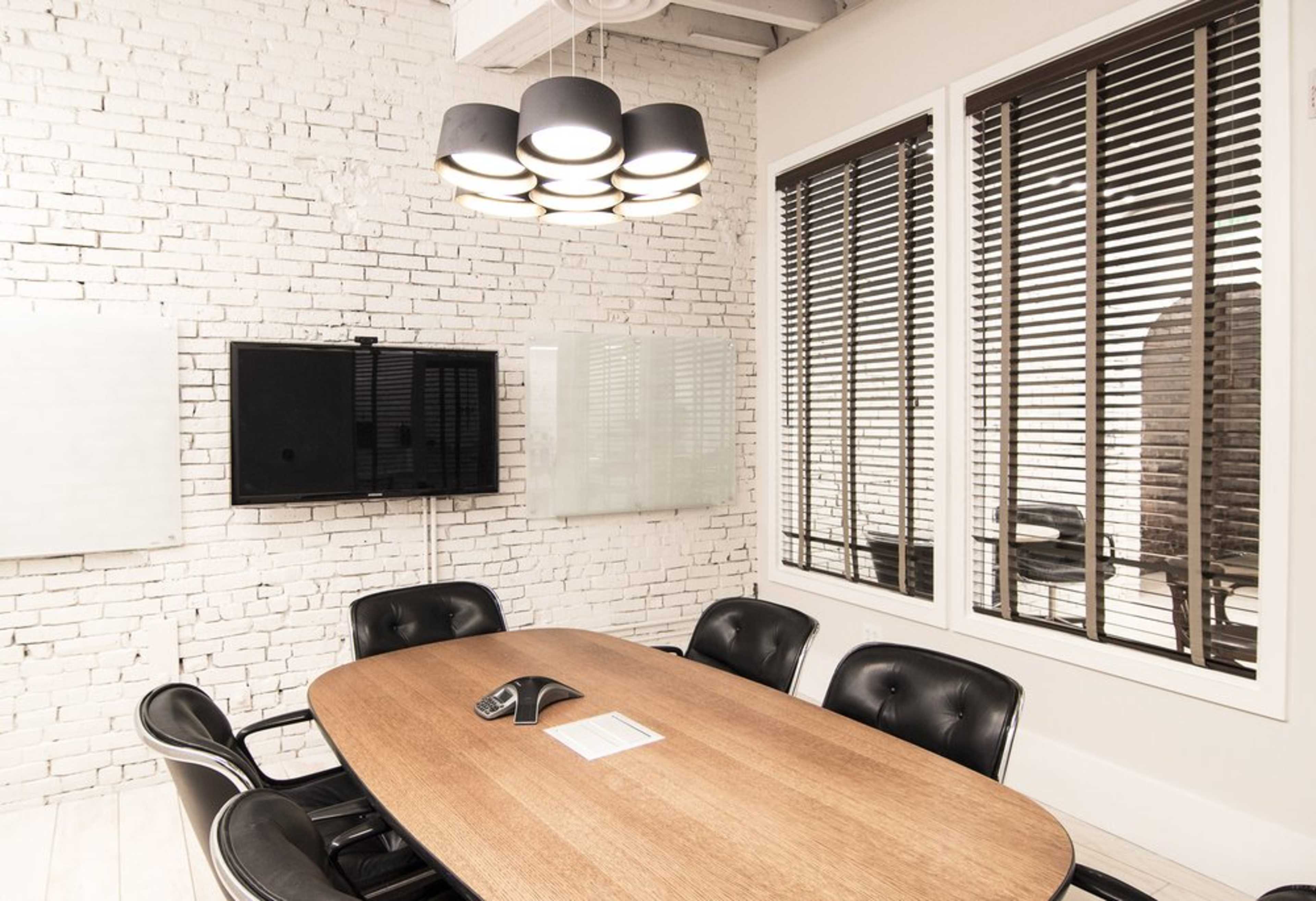 A modern conference room features a large oval wooden table surrounded by black leather chairs, with a television mounted on a white brick wall and large windows covered by blinds.