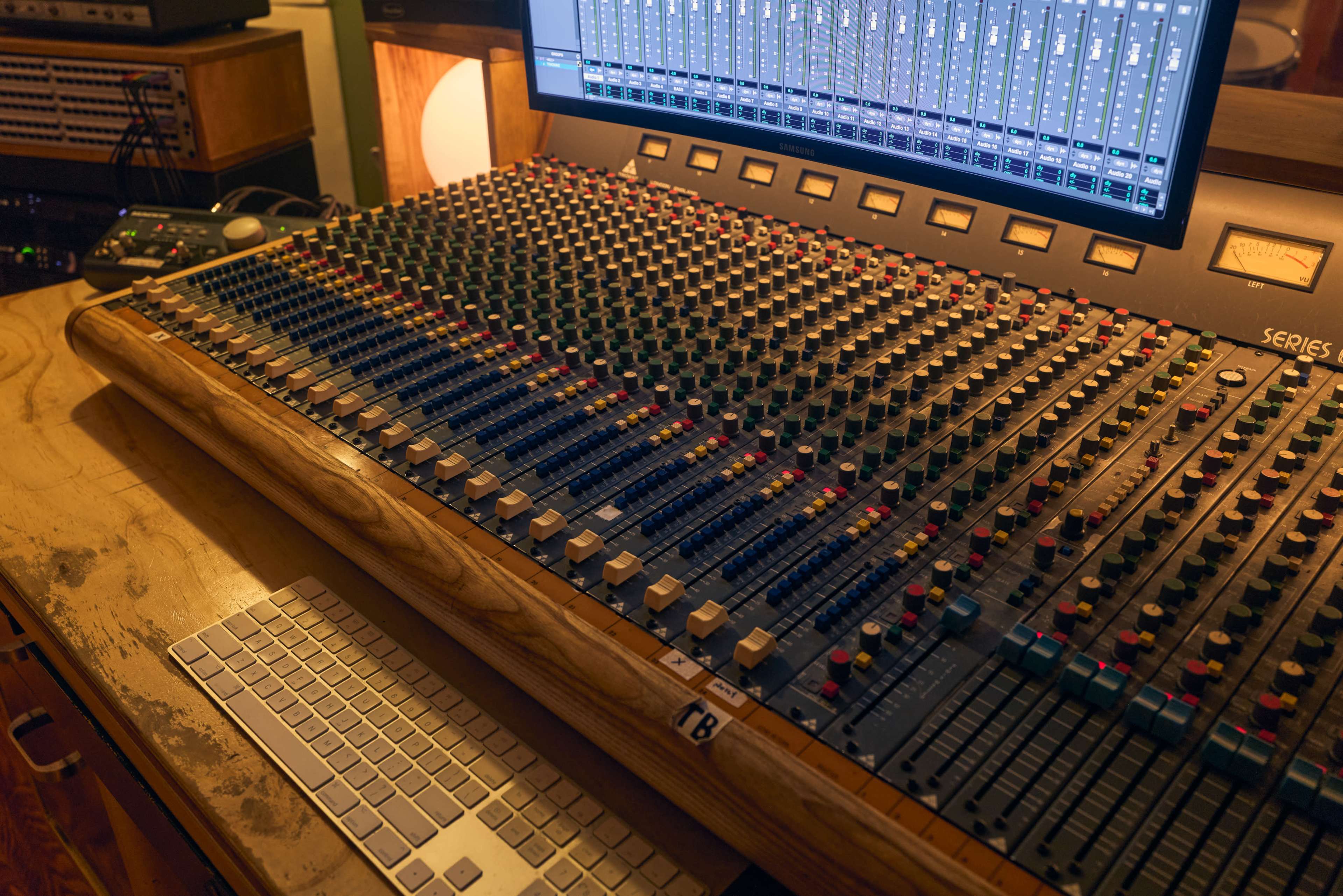 The image shows a large audio mixing console with numerous controls, faders, and a digital screen displaying tracks in a recording studio.