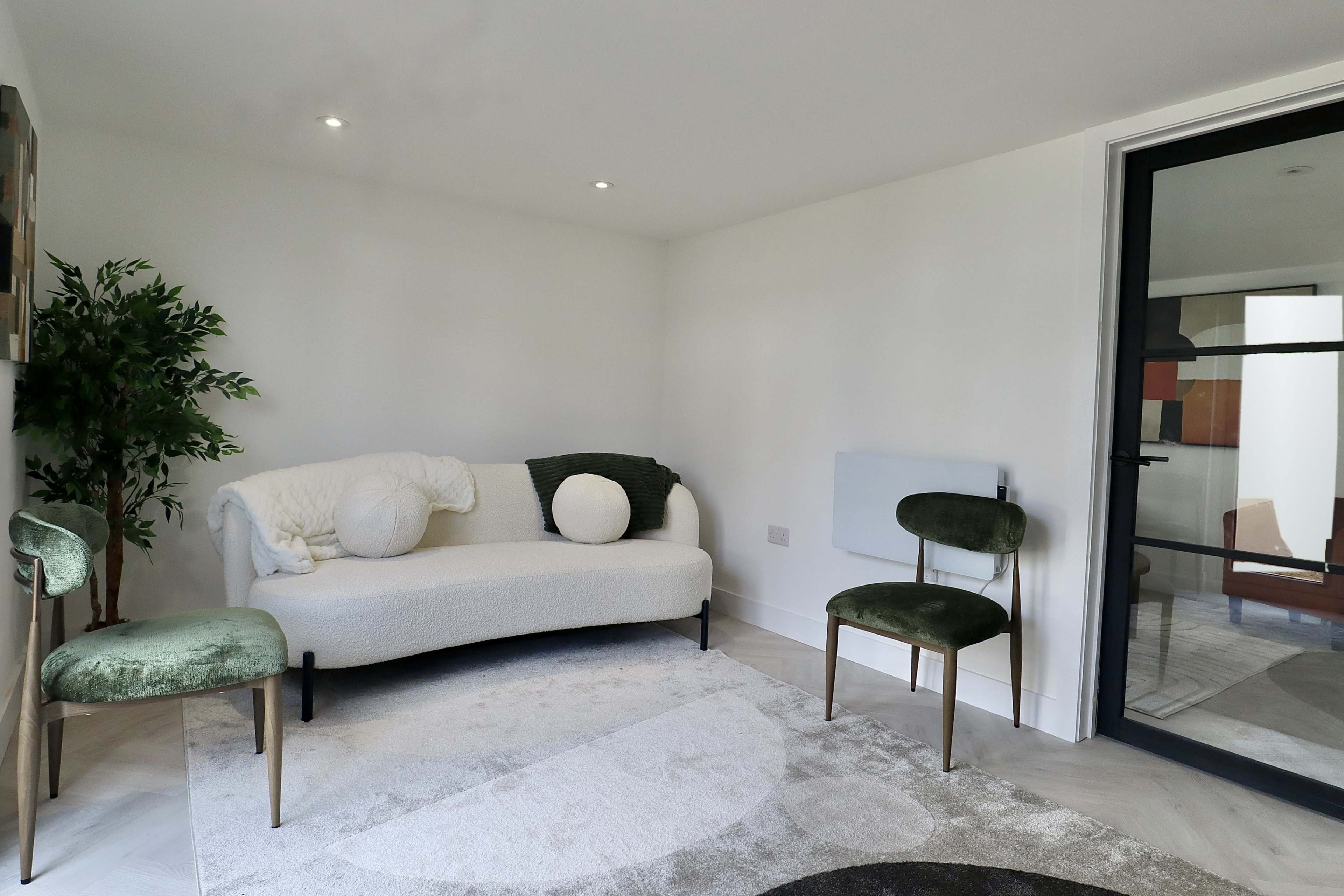 A small, modern living space features a white sofa, two green chairs, a plant, and a light-colored area rug.