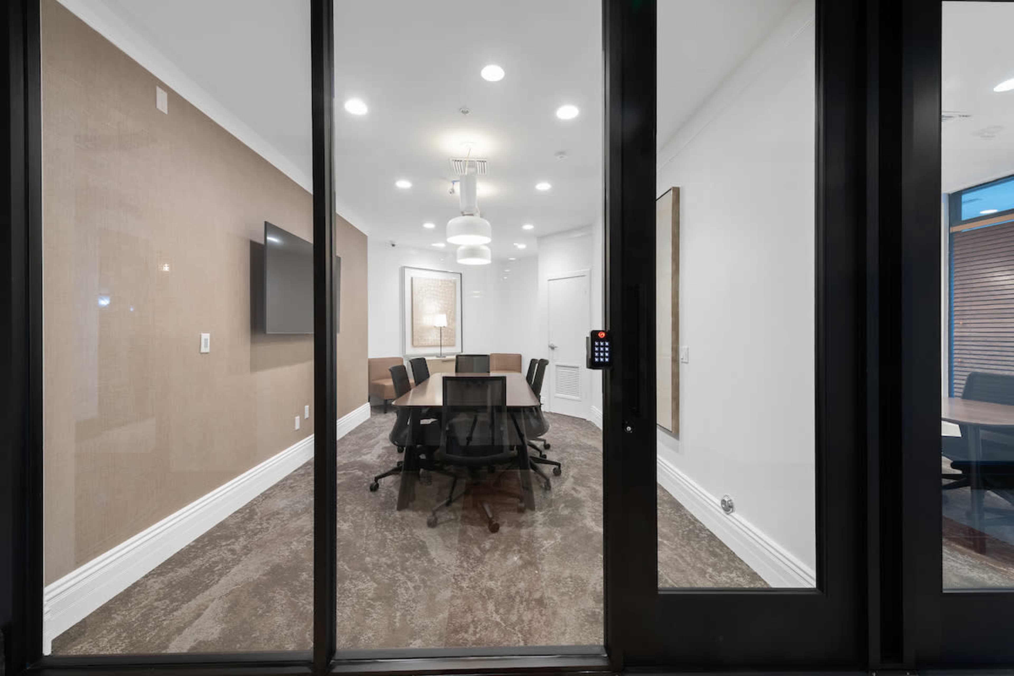 A glass-walled conference room features a large table surrounded by chairs, with a television mounted on the wall and a pendant light above.