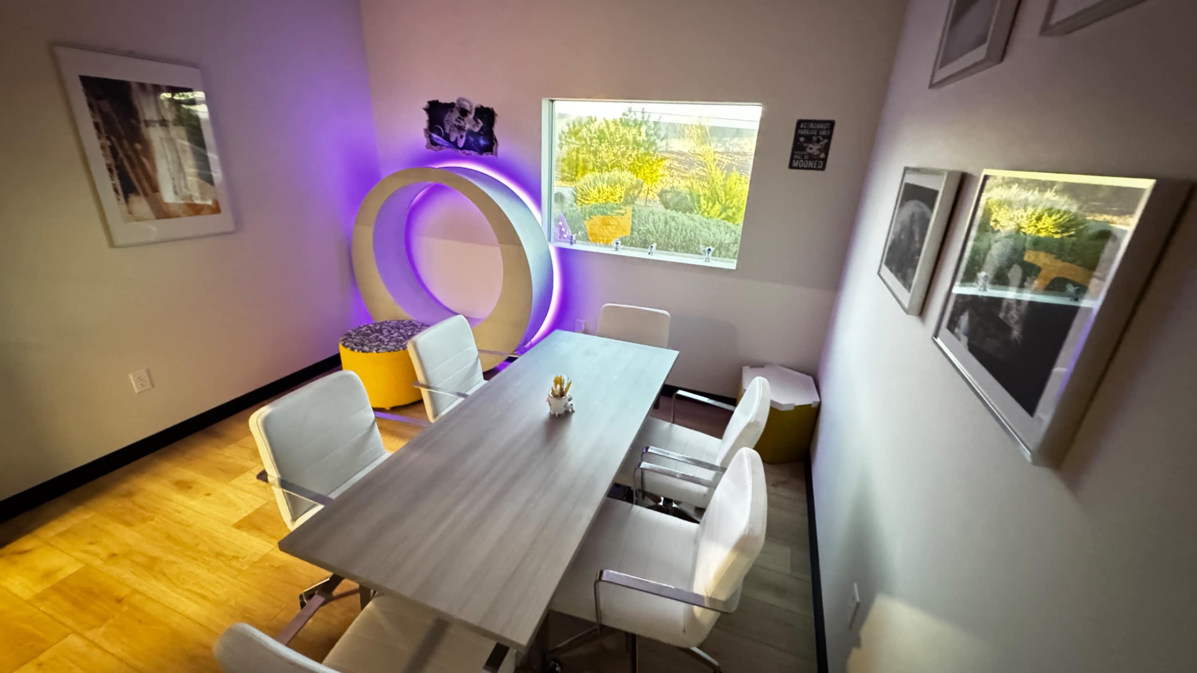 The image shows a modern meeting room featuring a long table surrounded by white chairs and a colorful decorative element with a circular shape illuminated in purple.