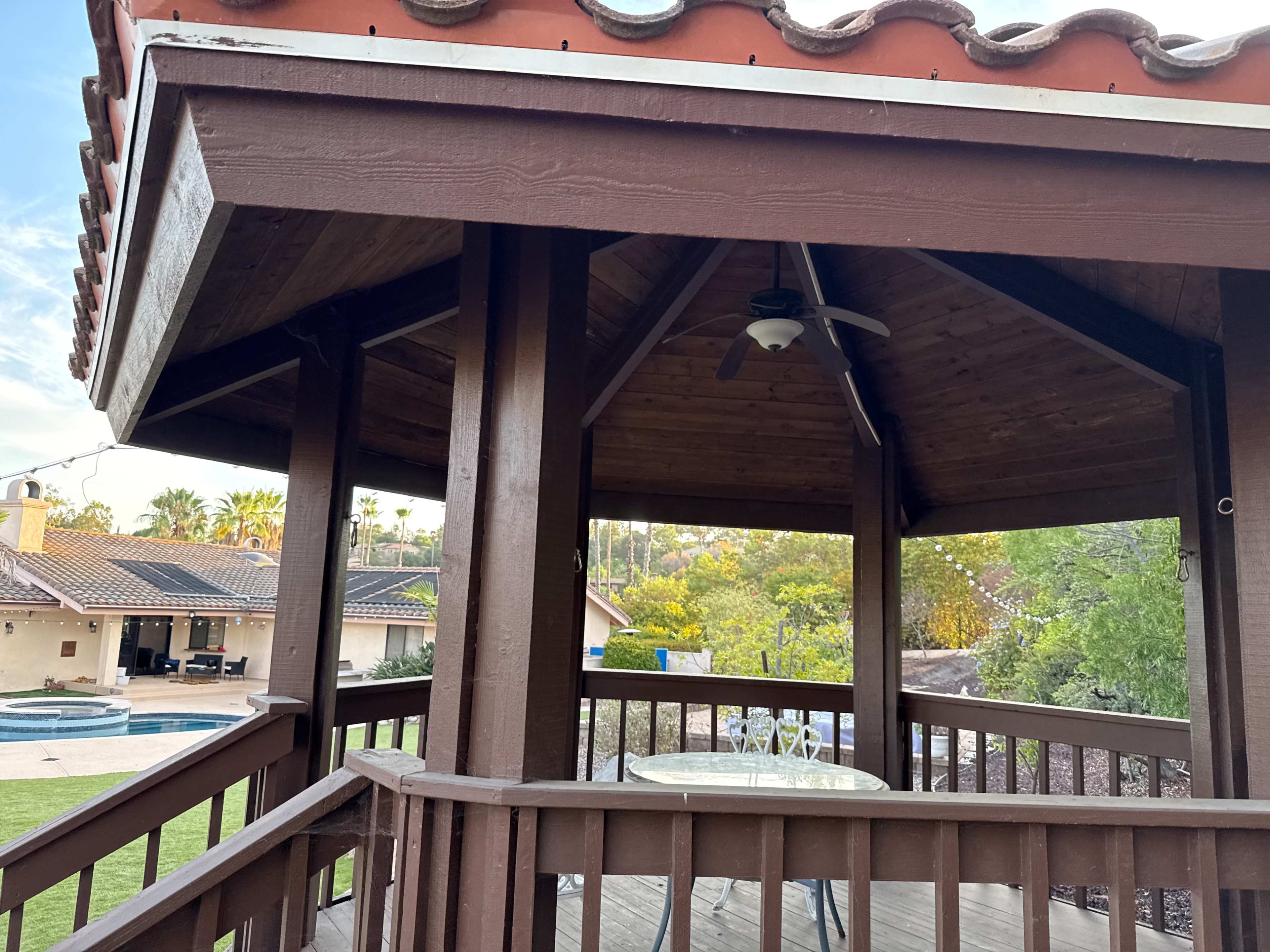The image shows a wooden gazebo with a table and chairs inside, overlooking a backyard with a swimming pool and surrounding greenery.