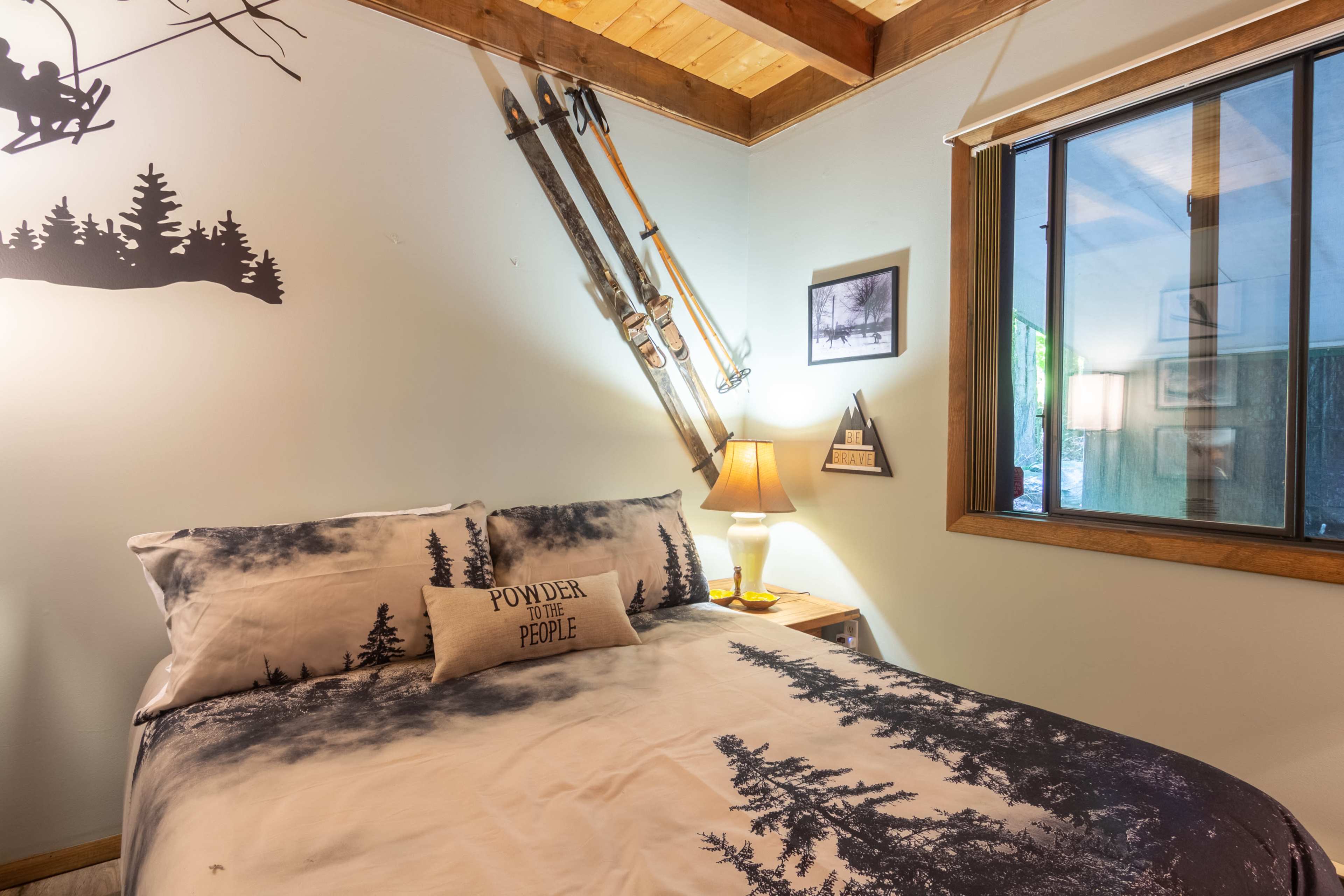 The cozy bedroom features a bed with a mountain-themed comforter, a bedside lamp, and a pair of vintage skis mounted on the wall.