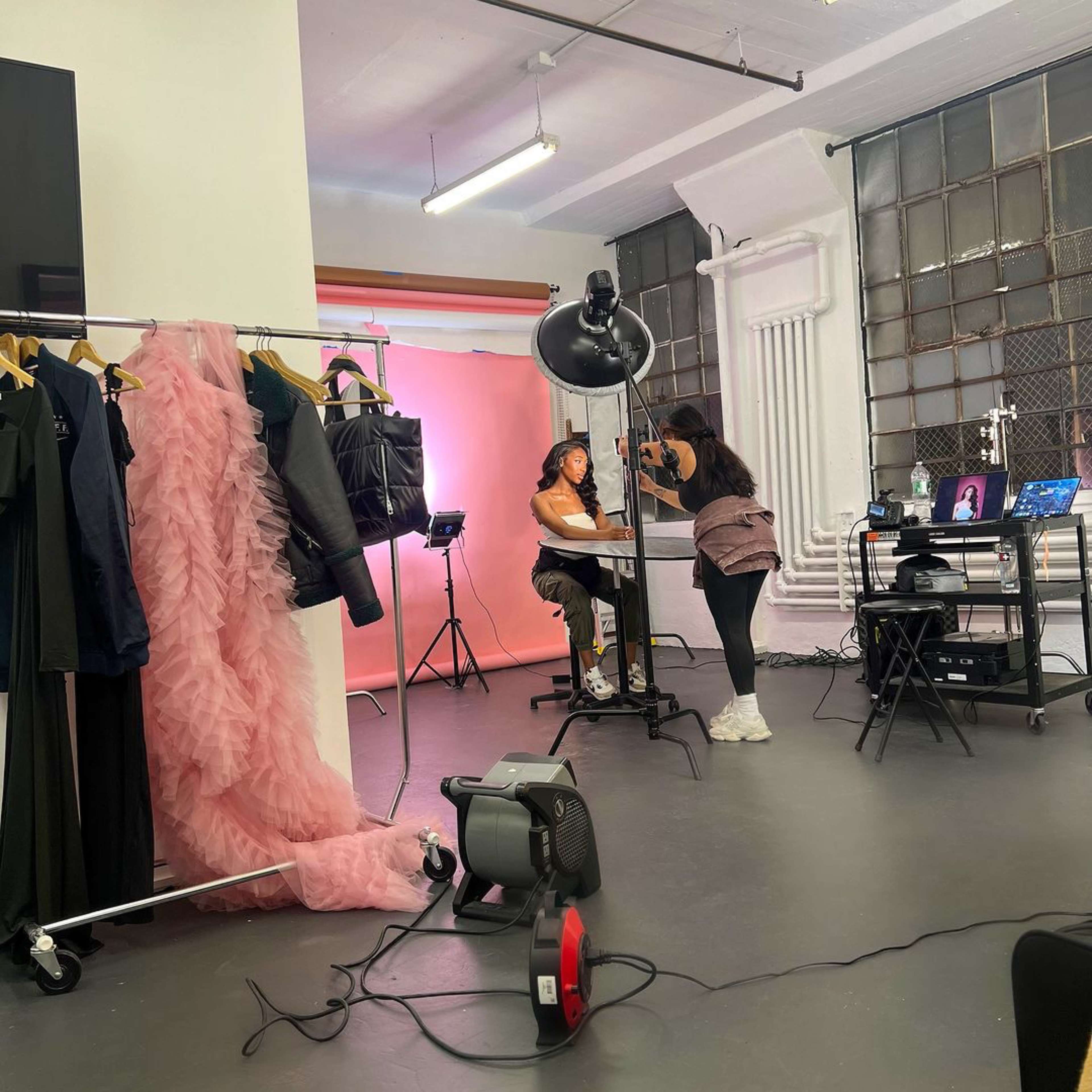 A photography studio setup features a model sitting at a table while a stylist adjusts her makeup, surrounded by clothing racks and a backdrop.