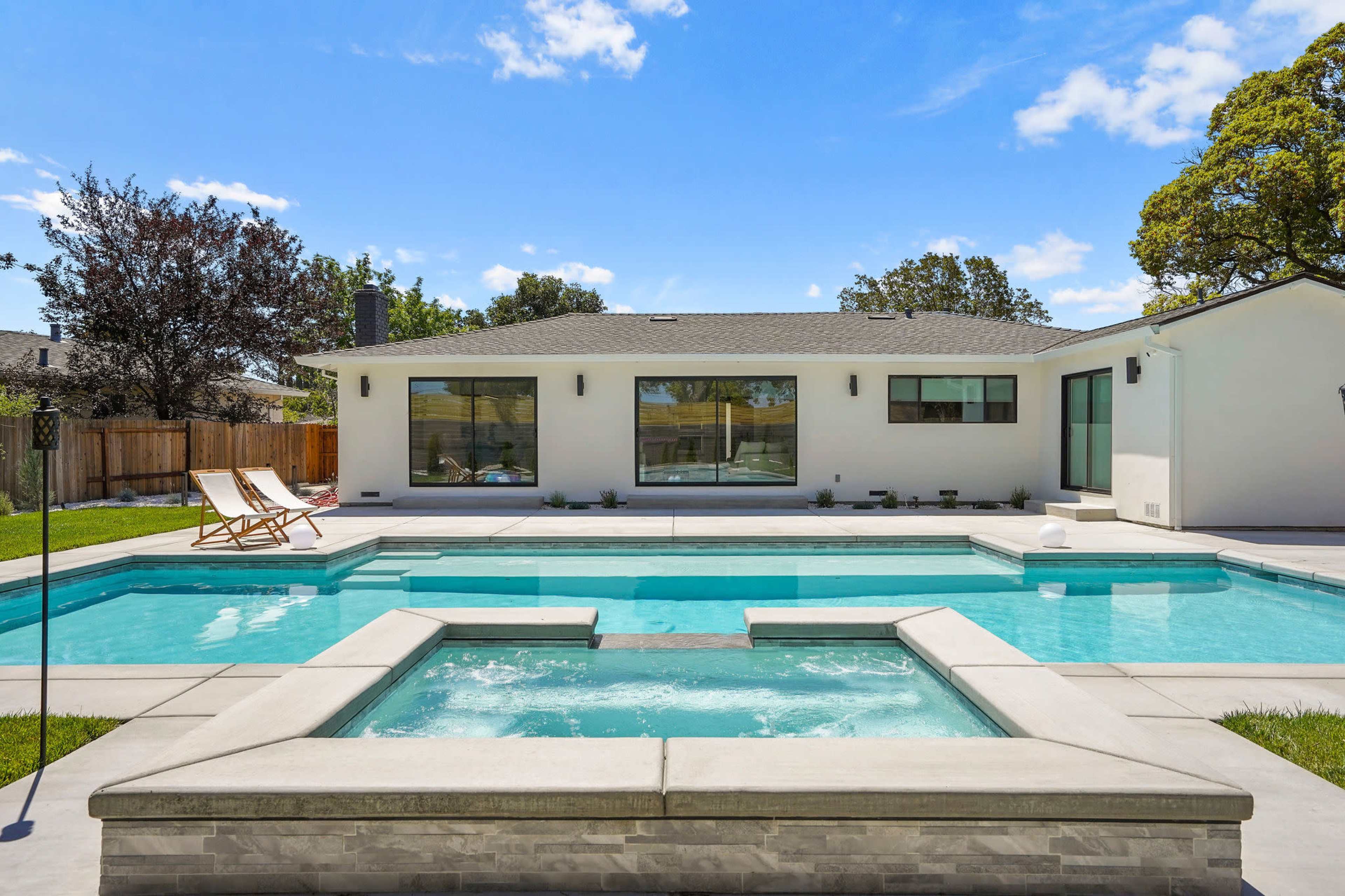 A modern home with large windows and a swimming pool featuring a spa area in the foreground surrounded by a landscaped yard.