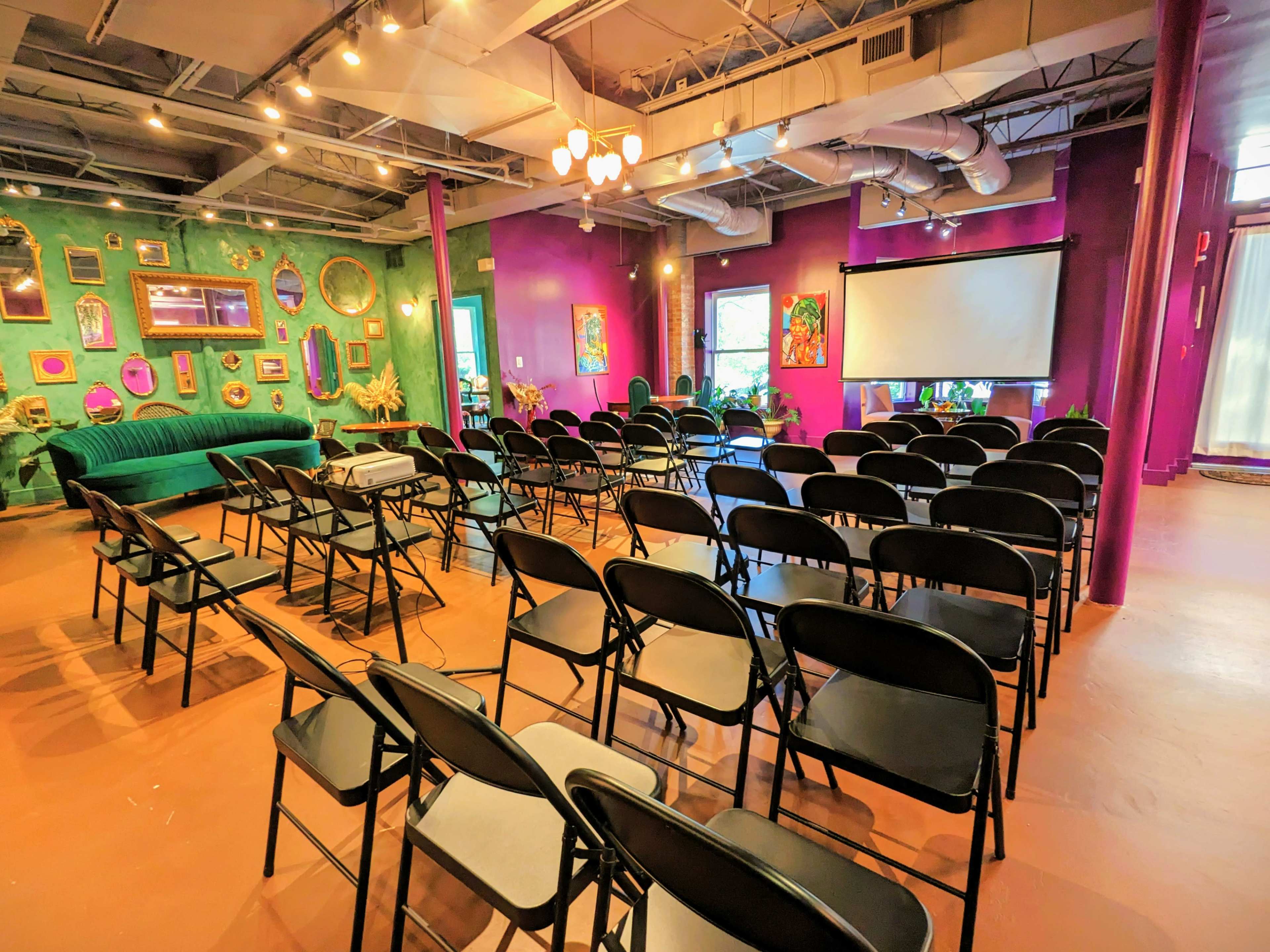 A room set up for an event, featuring rows of black chairs facing a projector screen, with a vibrant green and purple decor and a green couch against the wall.