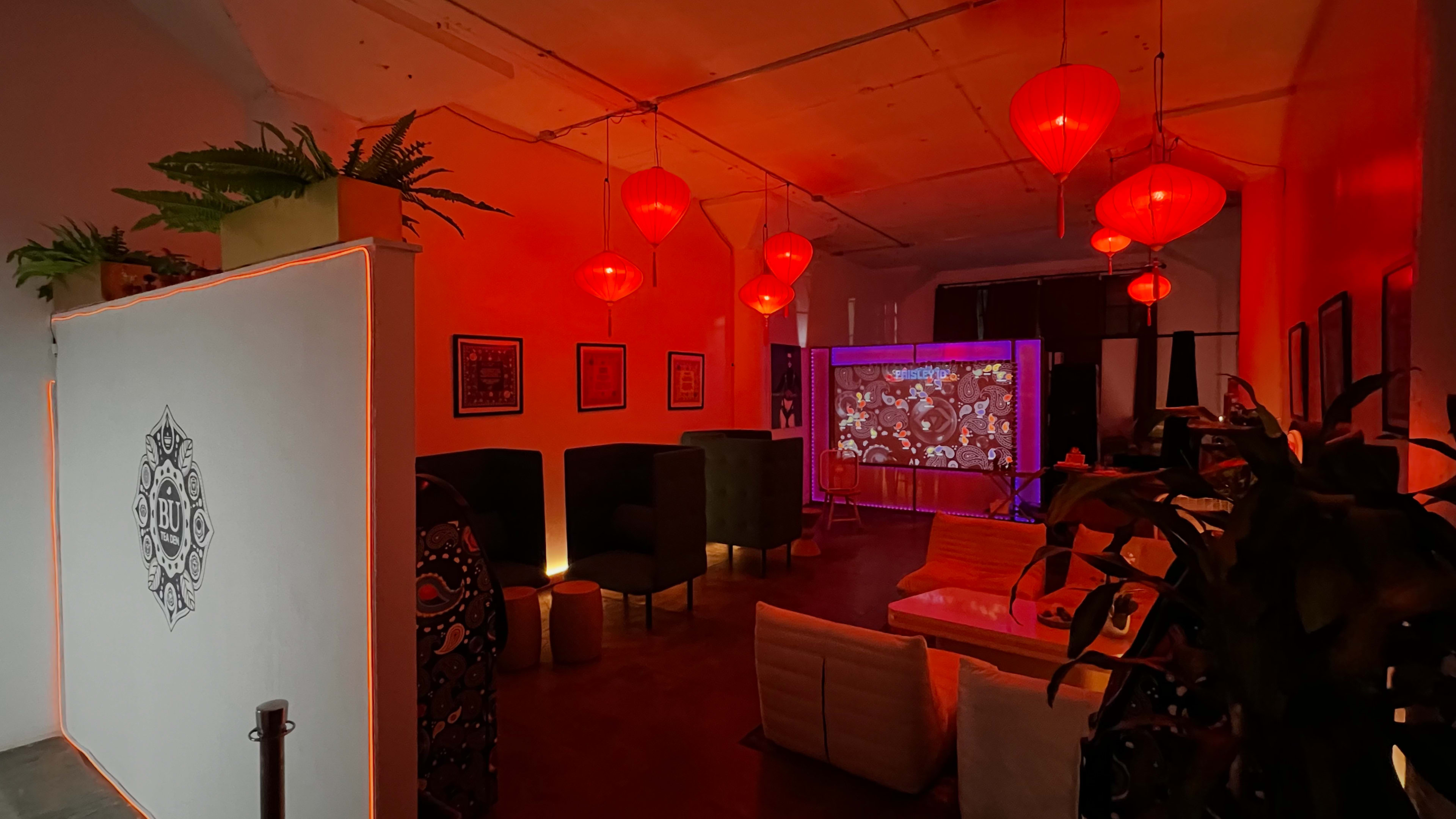 The image shows a dimly lit interior space with red lanterns hanging from the ceiling, seating areas with dark-colored furniture, and a large projection screen displaying colorful visuals.