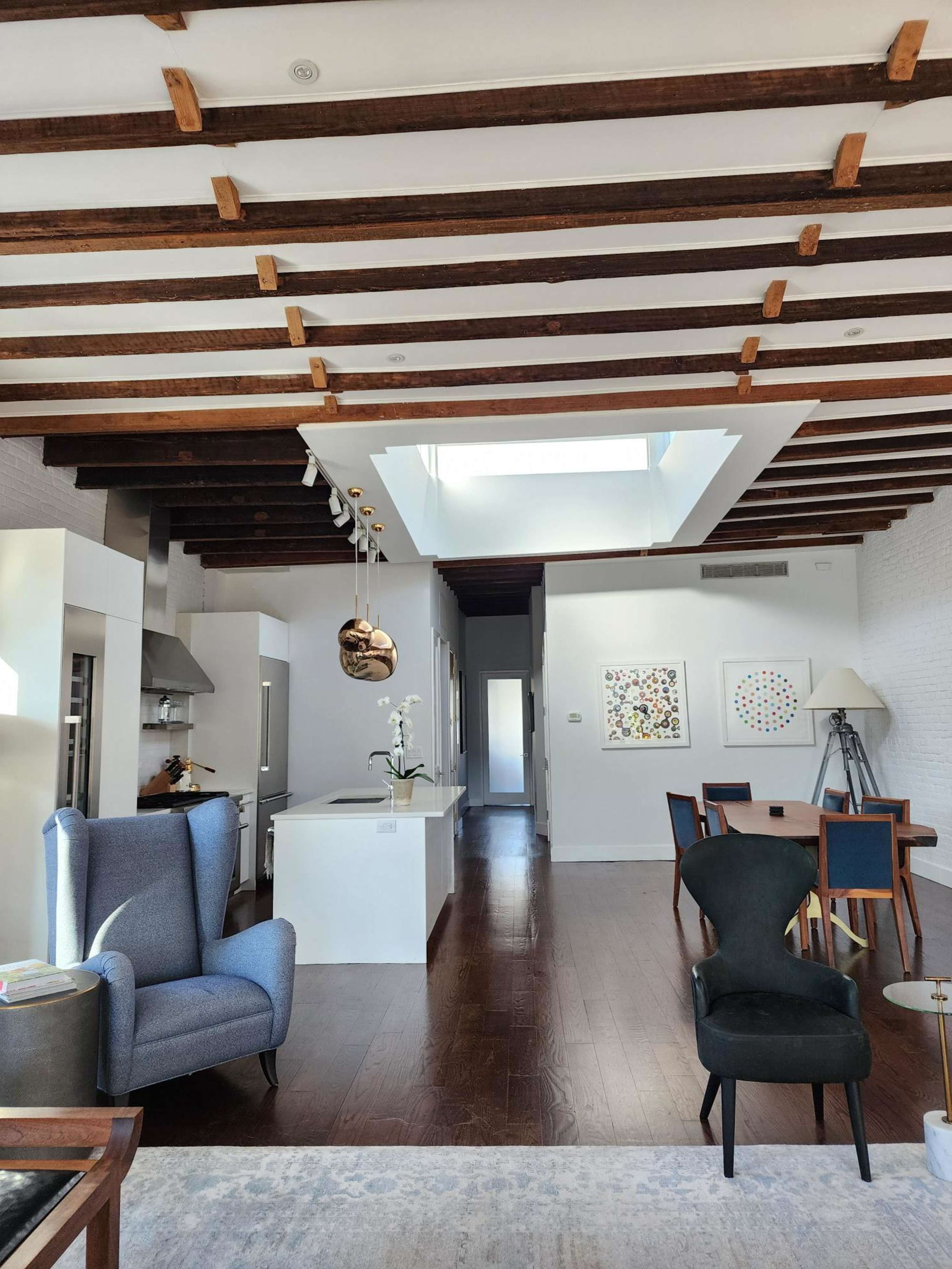 A modern open-concept living space with wooden beams, a kitchen area, a dining table, and a skylight.