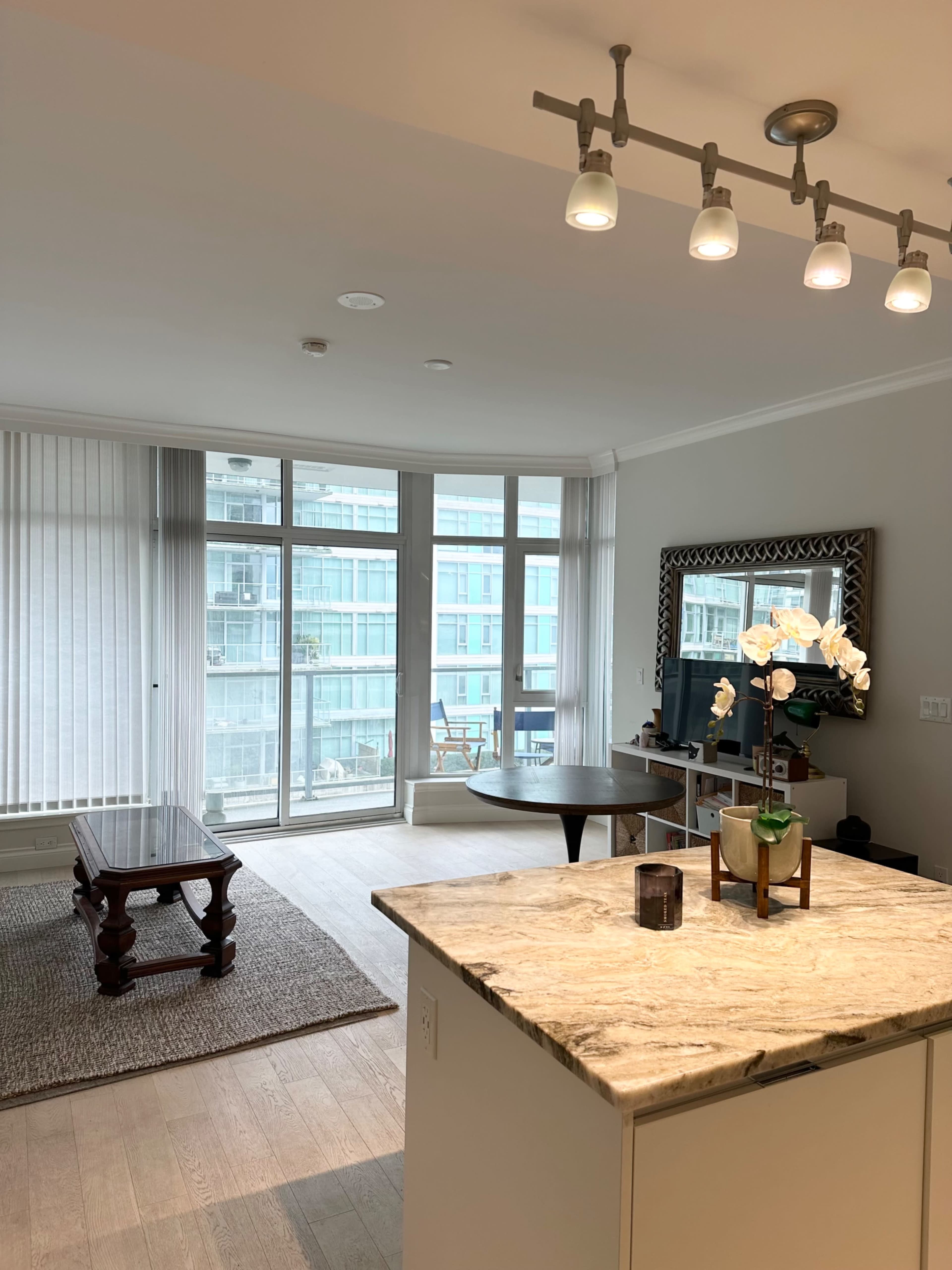The image shows a modern apartment interior featuring a kitchen island with a marble countertop, a dining area, and large windows with a view of a balcony.