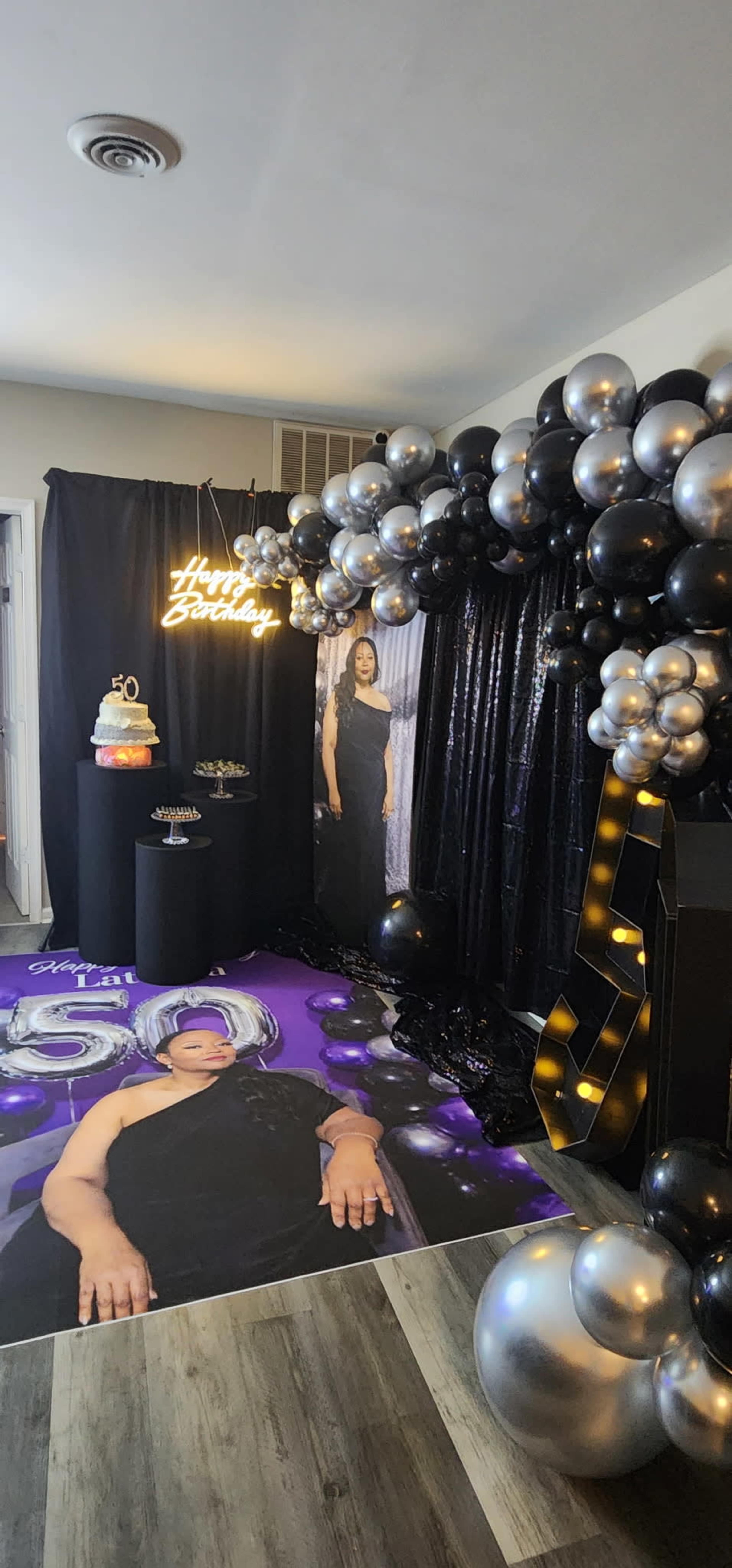 The image shows a decorated birthday party scene featuring a black and silver balloon arch, a large photo display of the birthday person, and a cake on a table.