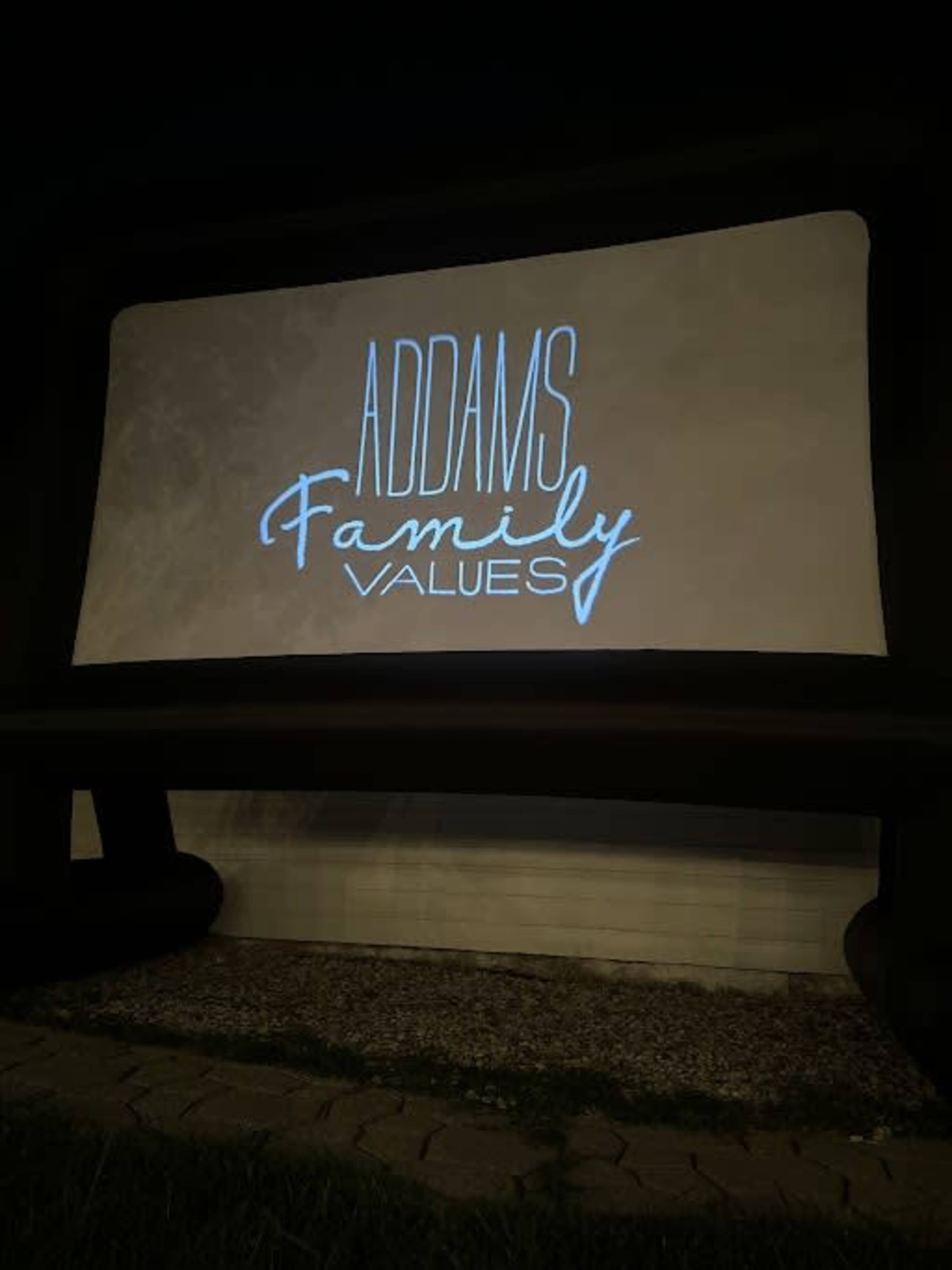 A projection screen displaying the title "Addams Family Values" is set up on an outdoor backdrop.