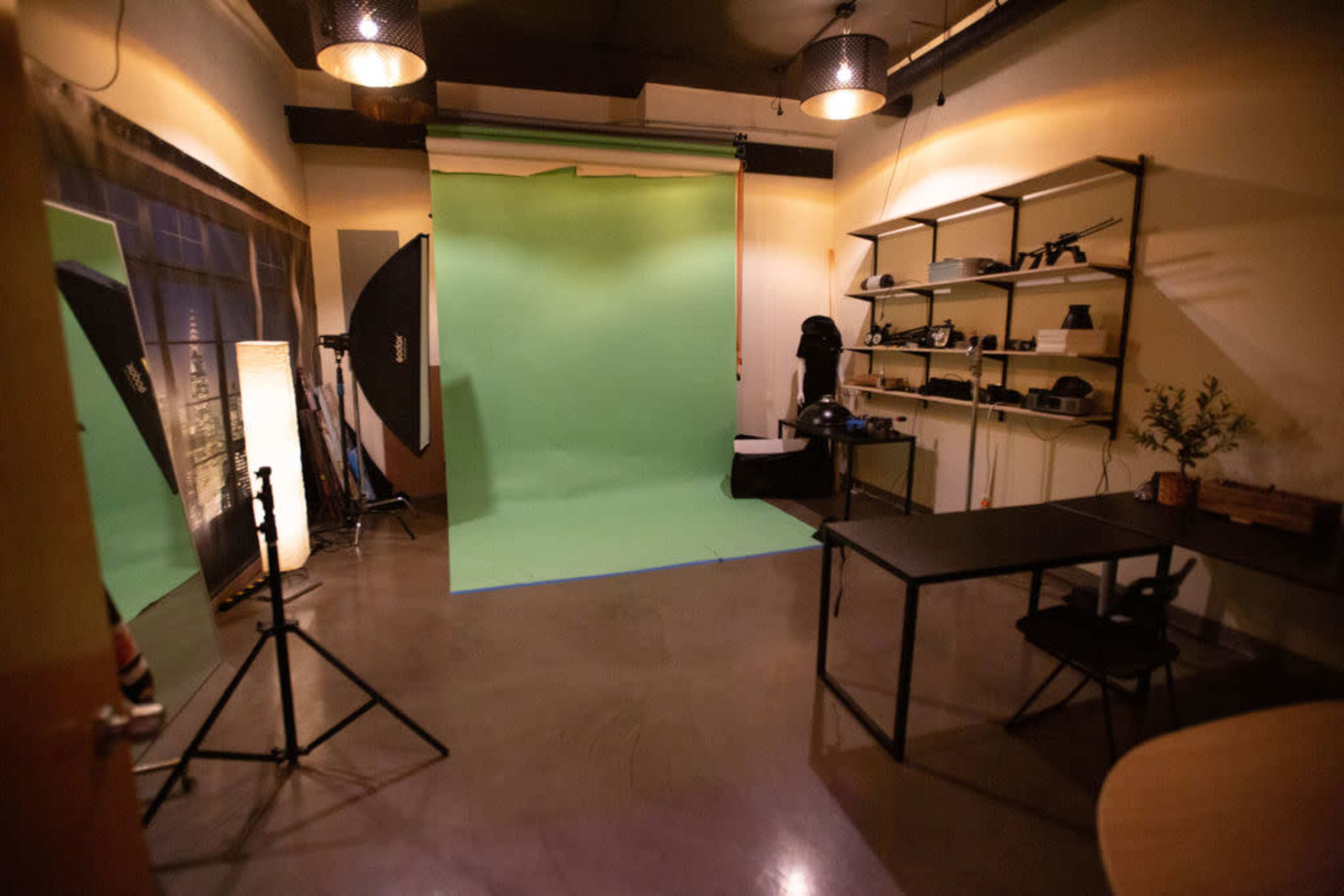 The image shows a photography studio featuring a green backdrop, lighting equipment, a black table with a chair, and shelves filled with camera accessories.