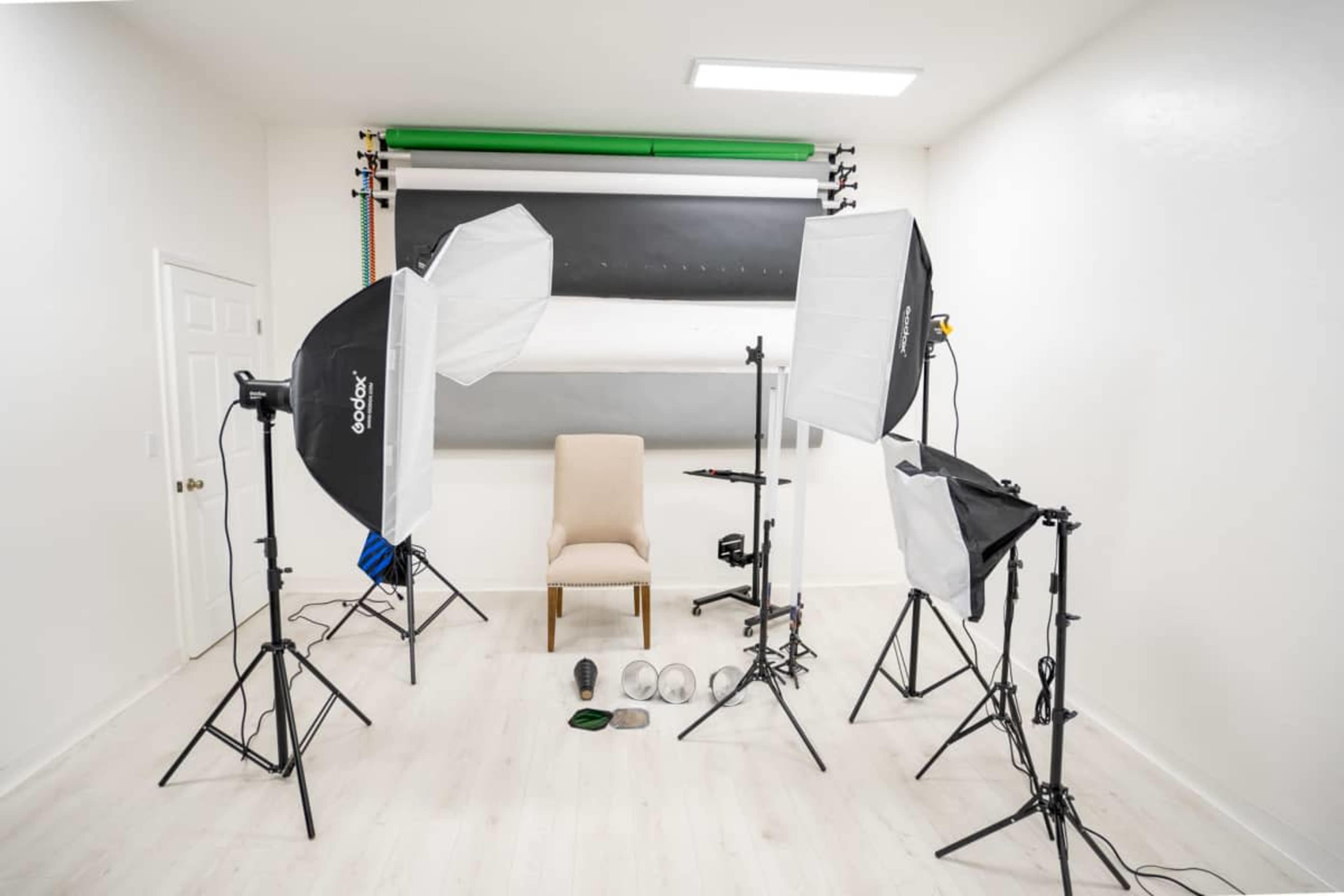 The image shows a photography studio setup with two softbox lights, a backdrop stand with a gray backdrop, an empty chair in the center, and various props on the floor.