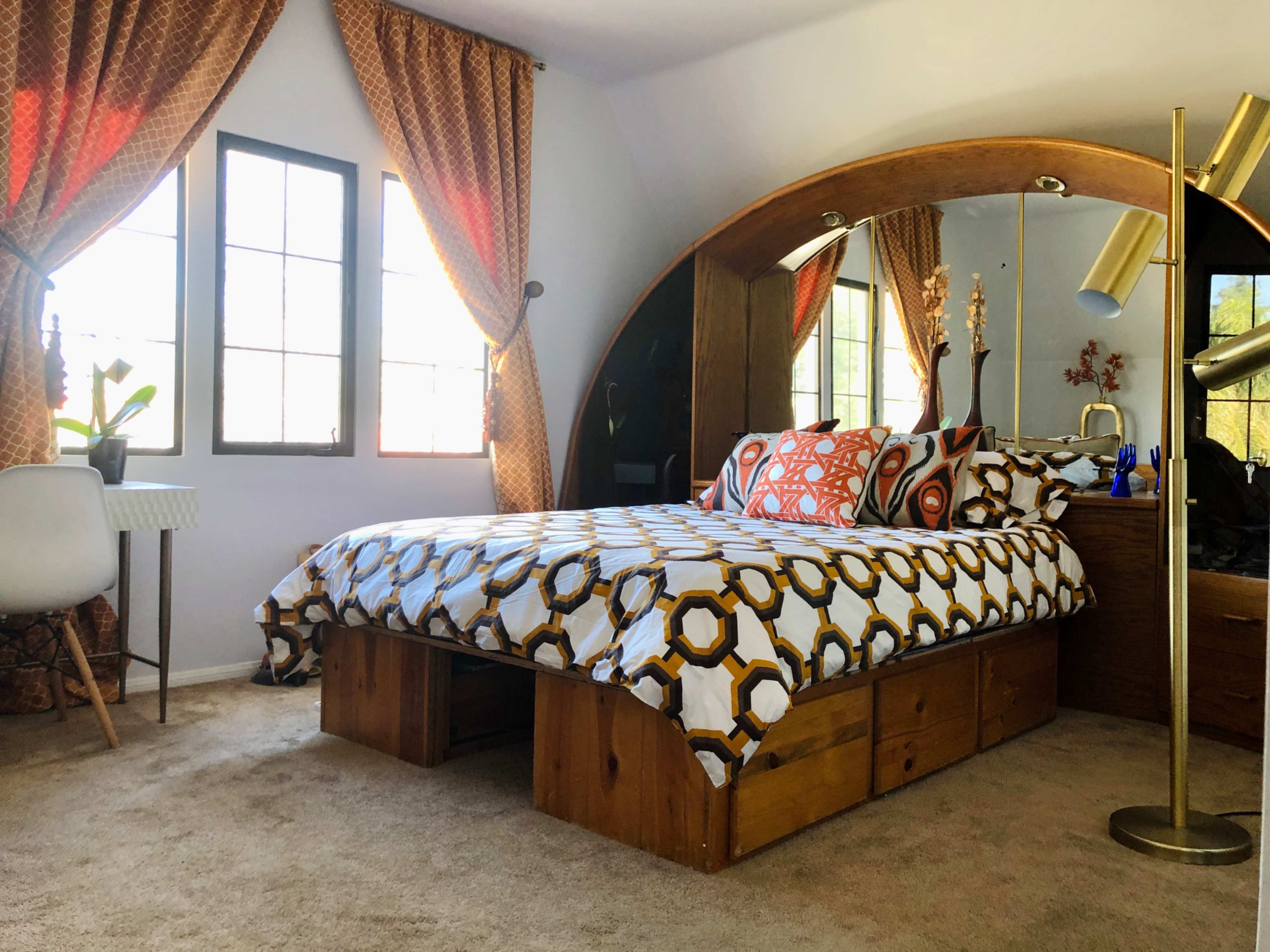 A bedroom features a bed with patterned bedding, a wooden headboard, two windows with drapes, and a mirror reflecting the room's decor.