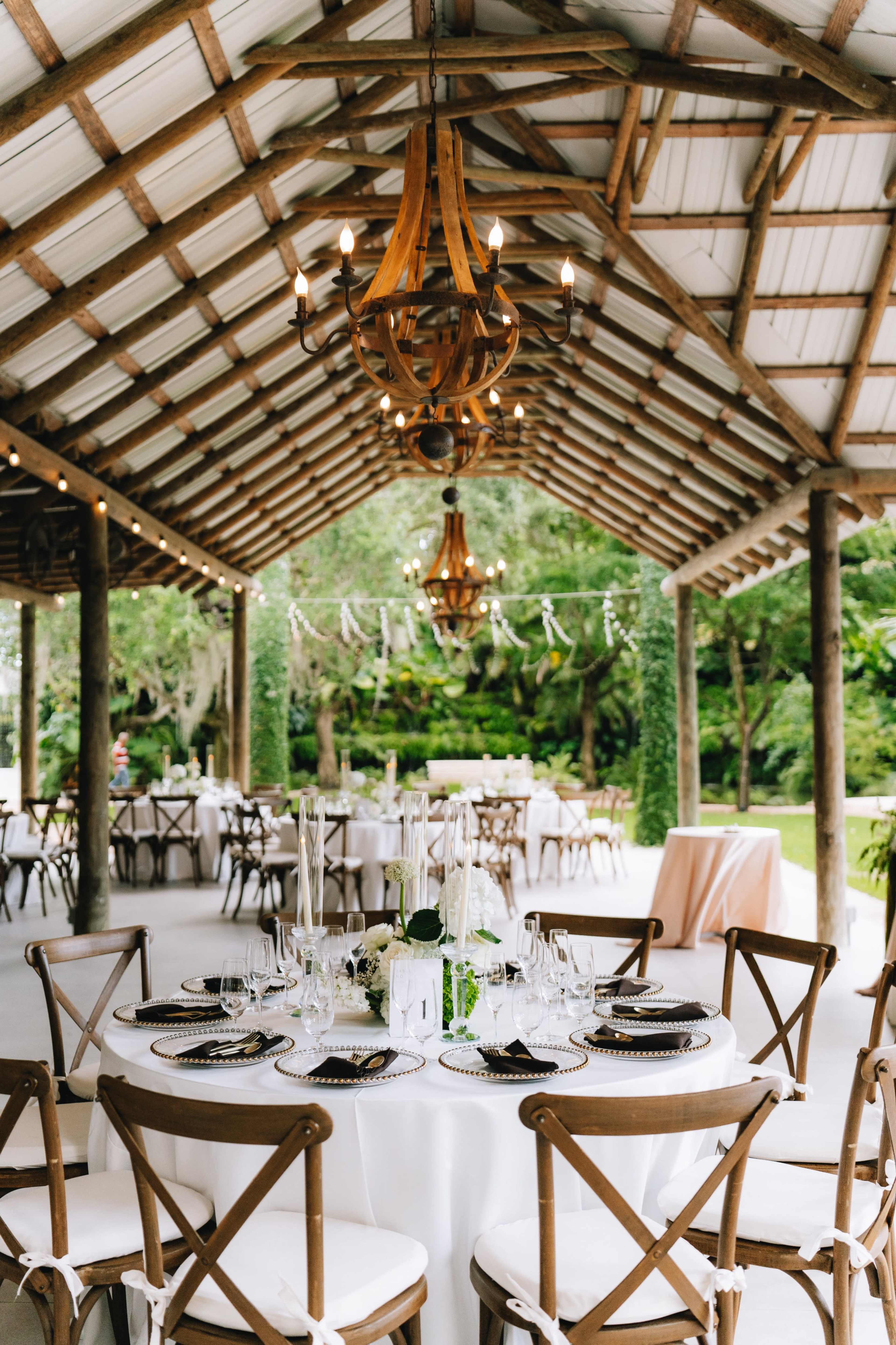 A spacious outdoor dining area features elegantly set tables under a rustic wooden structure with chandeliers and greenery in the background.
