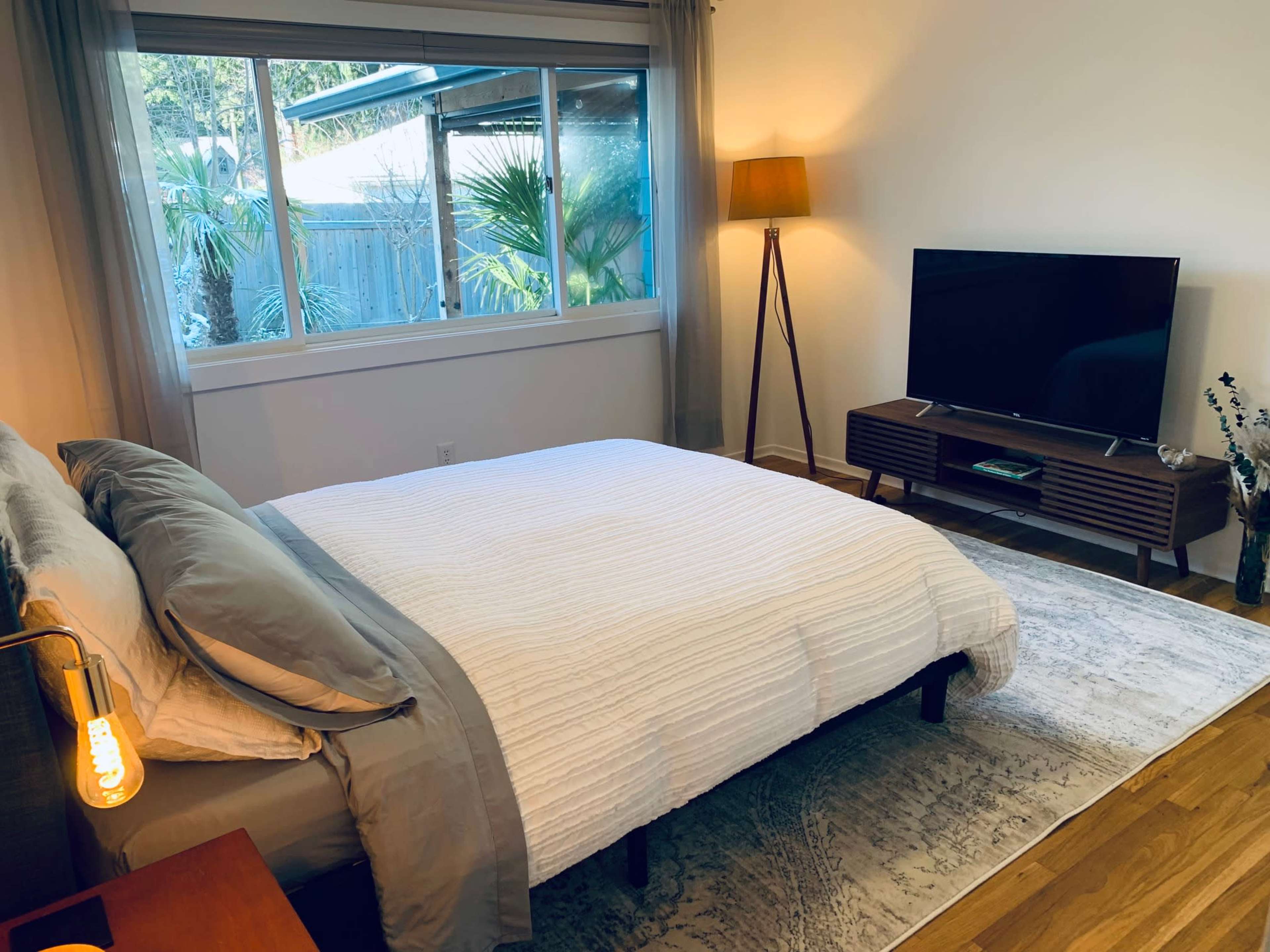 A well-lit bedroom featuring a bed with white bedding, a standing lamp, a television on a wooden console, and a large window with a view of greenery outside.