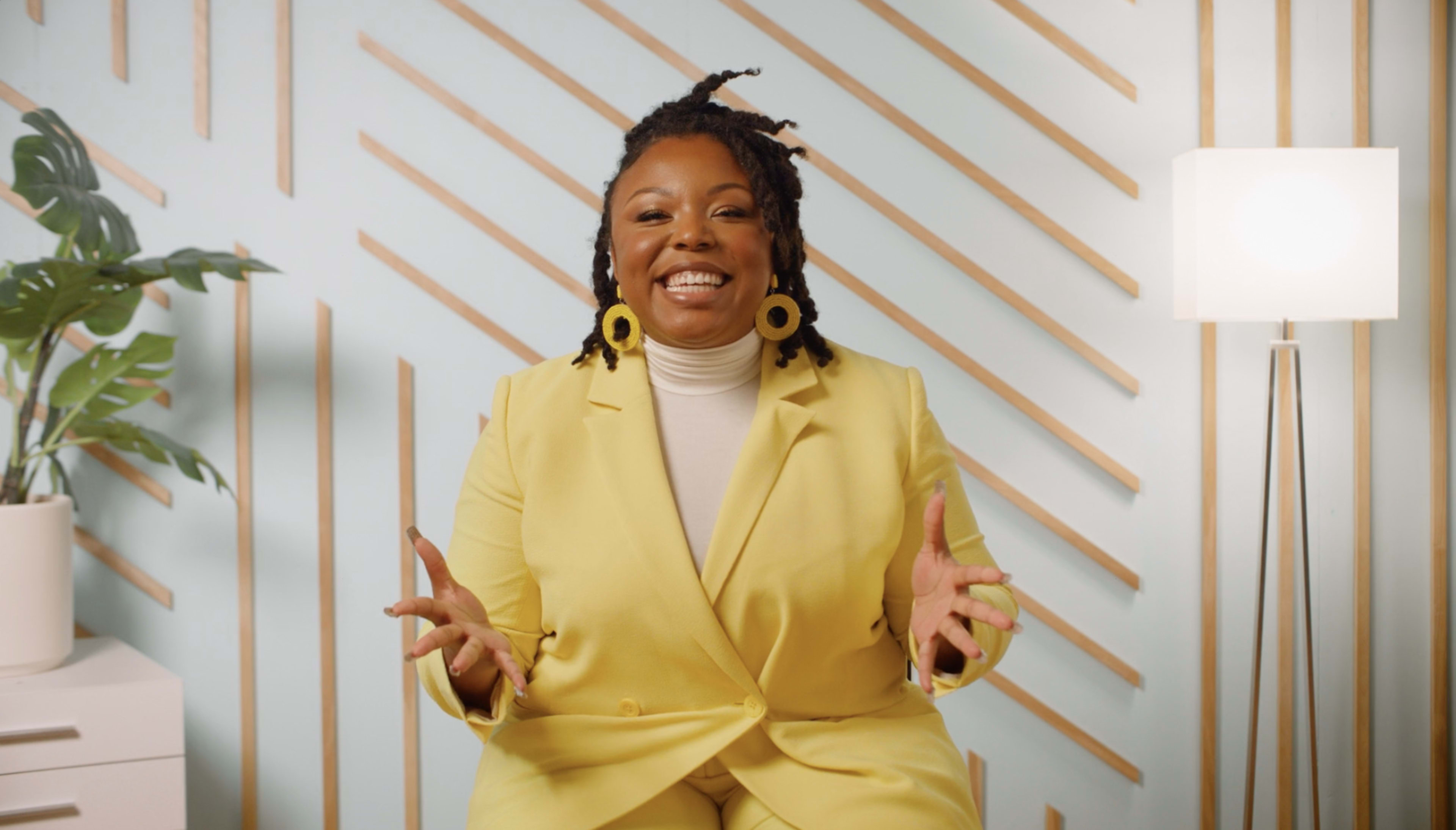 A woman in a yellow suit sits in front of a light blue wall with diagonal wooden slats, smiling and gesturing with her hands.