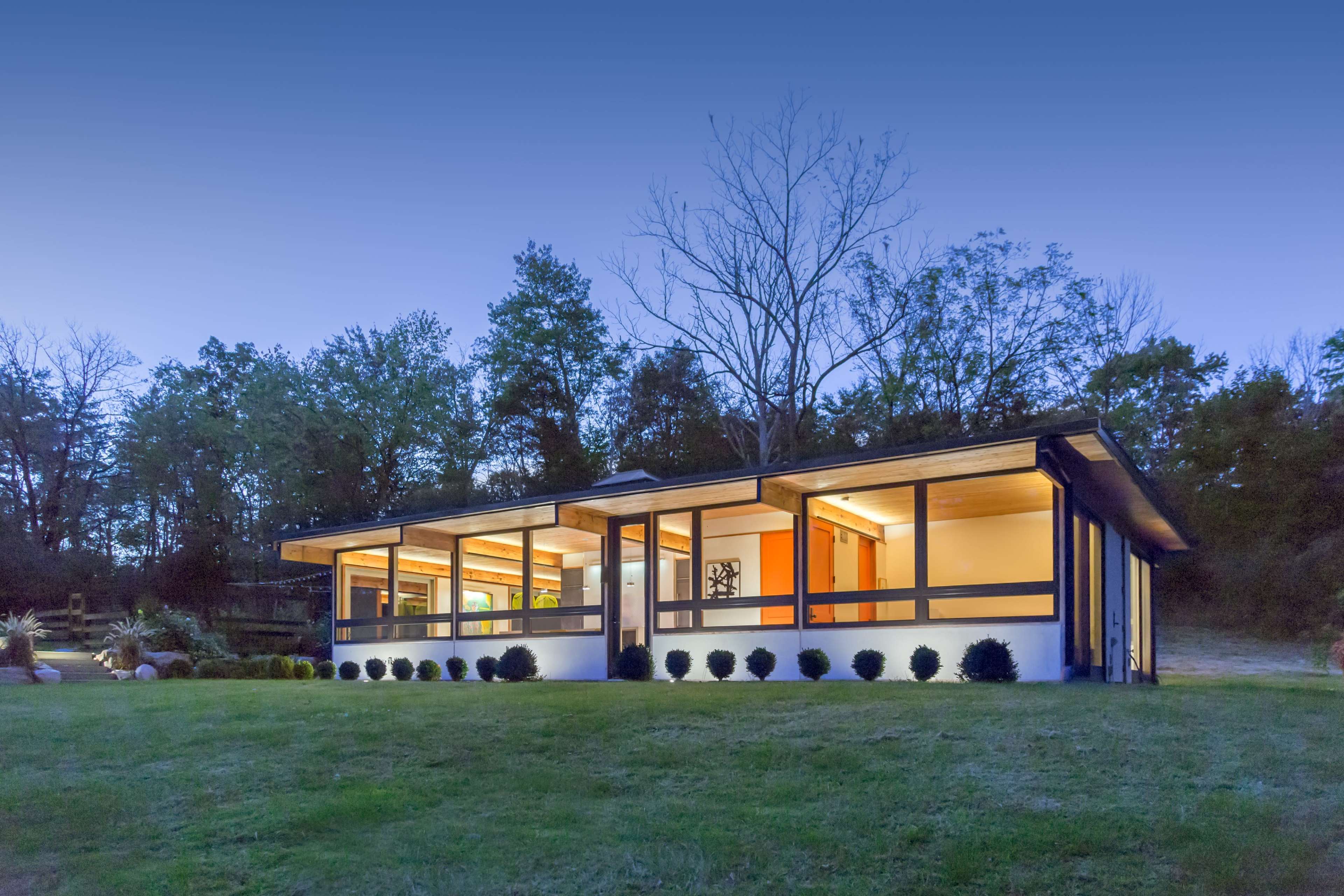 A modern house with large windows is set against a backdrop of trees in the evening light.