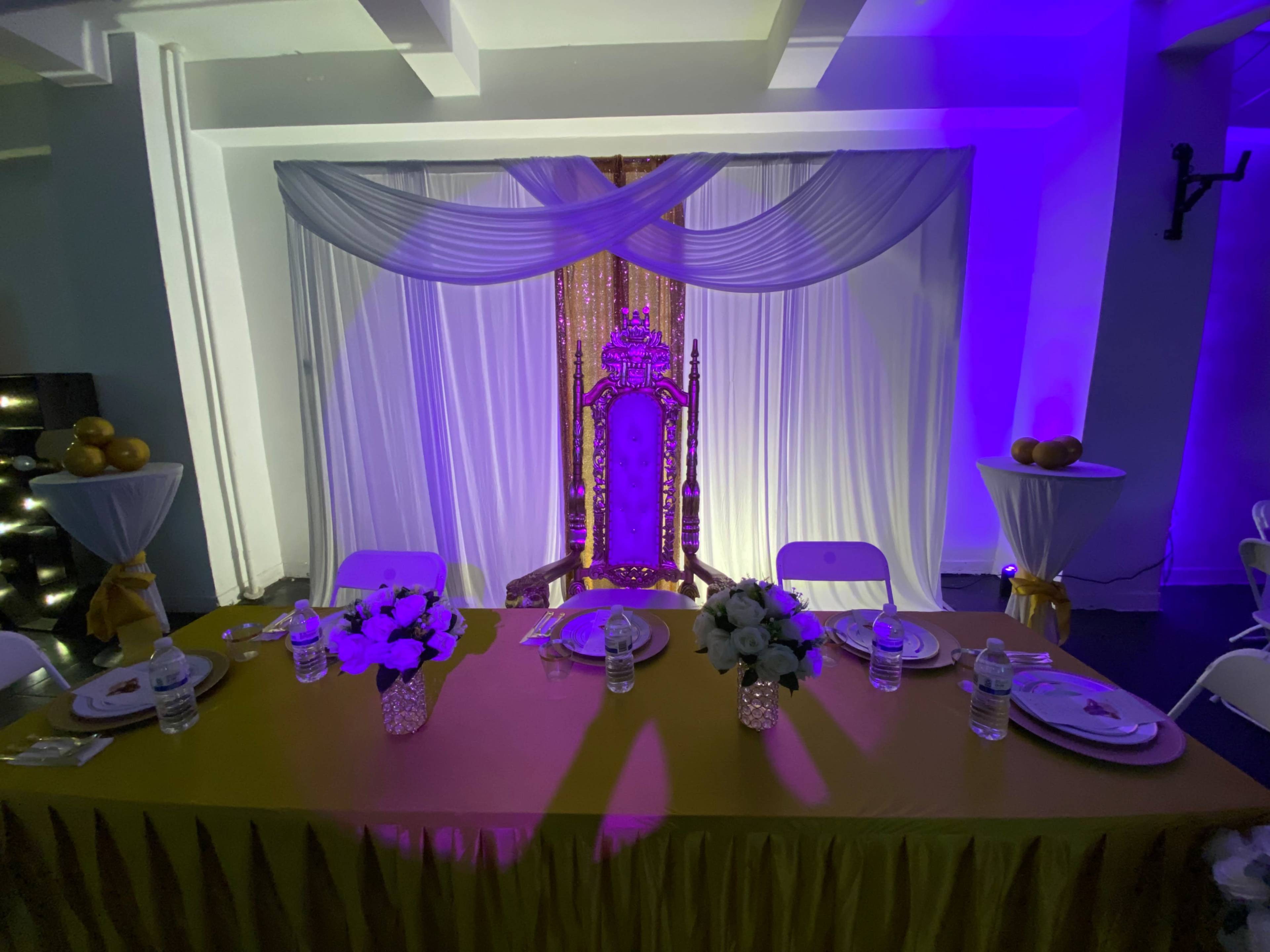 The scene features a decorated table set for an event, with a vibrant throne backdrop, draped in white fabric and illuminated by purple lighting.
